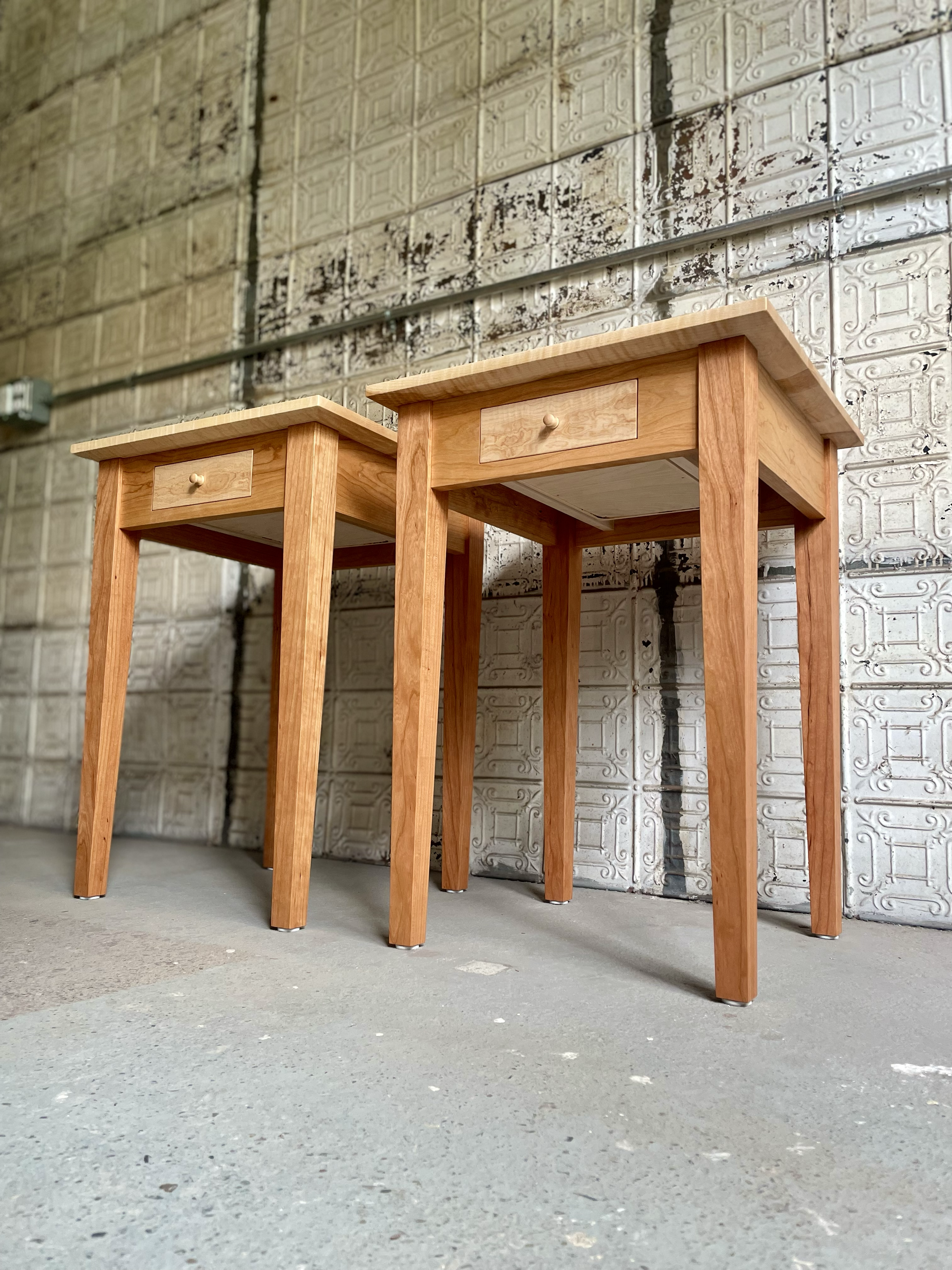 Two wooden side tables with small drawers, positioned against a vintage metal wall in a warehouse-like setting.