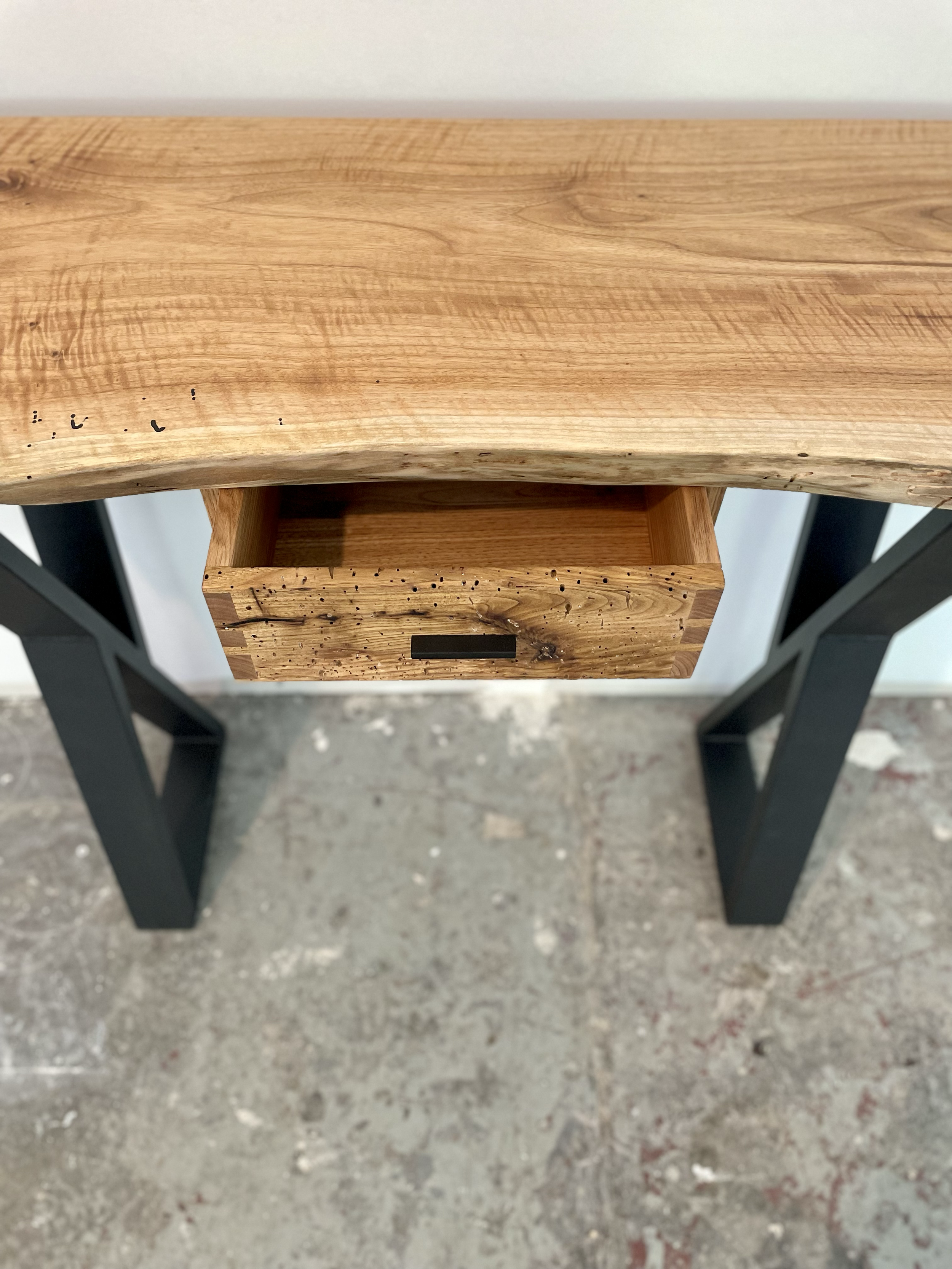 Top view of a wooden desk with a single open drawer, supported by black metal legs, on a concrete floor.