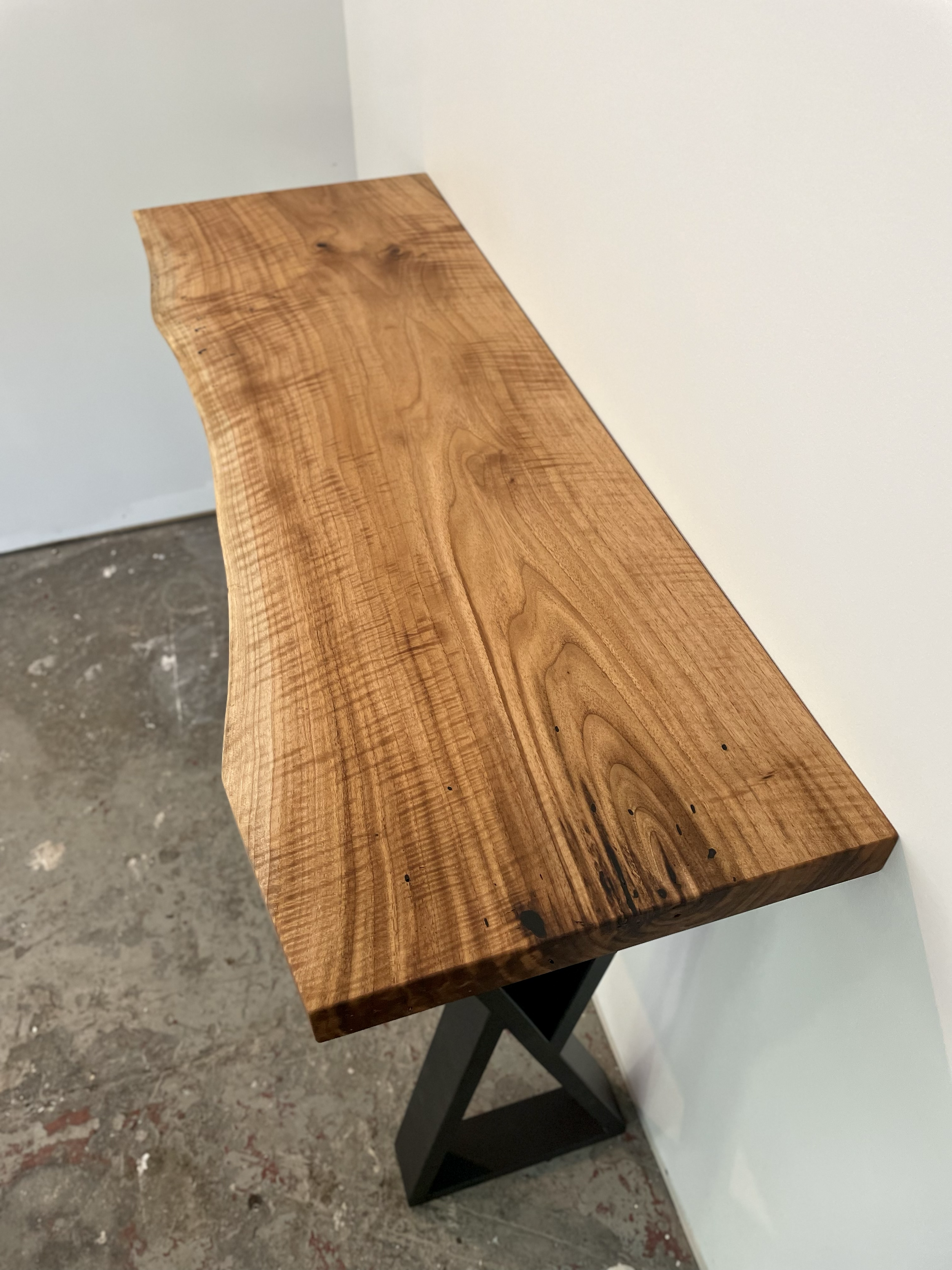 A wooden table with a live edge, mounted on black metal trestle legs, placed against a white wall.