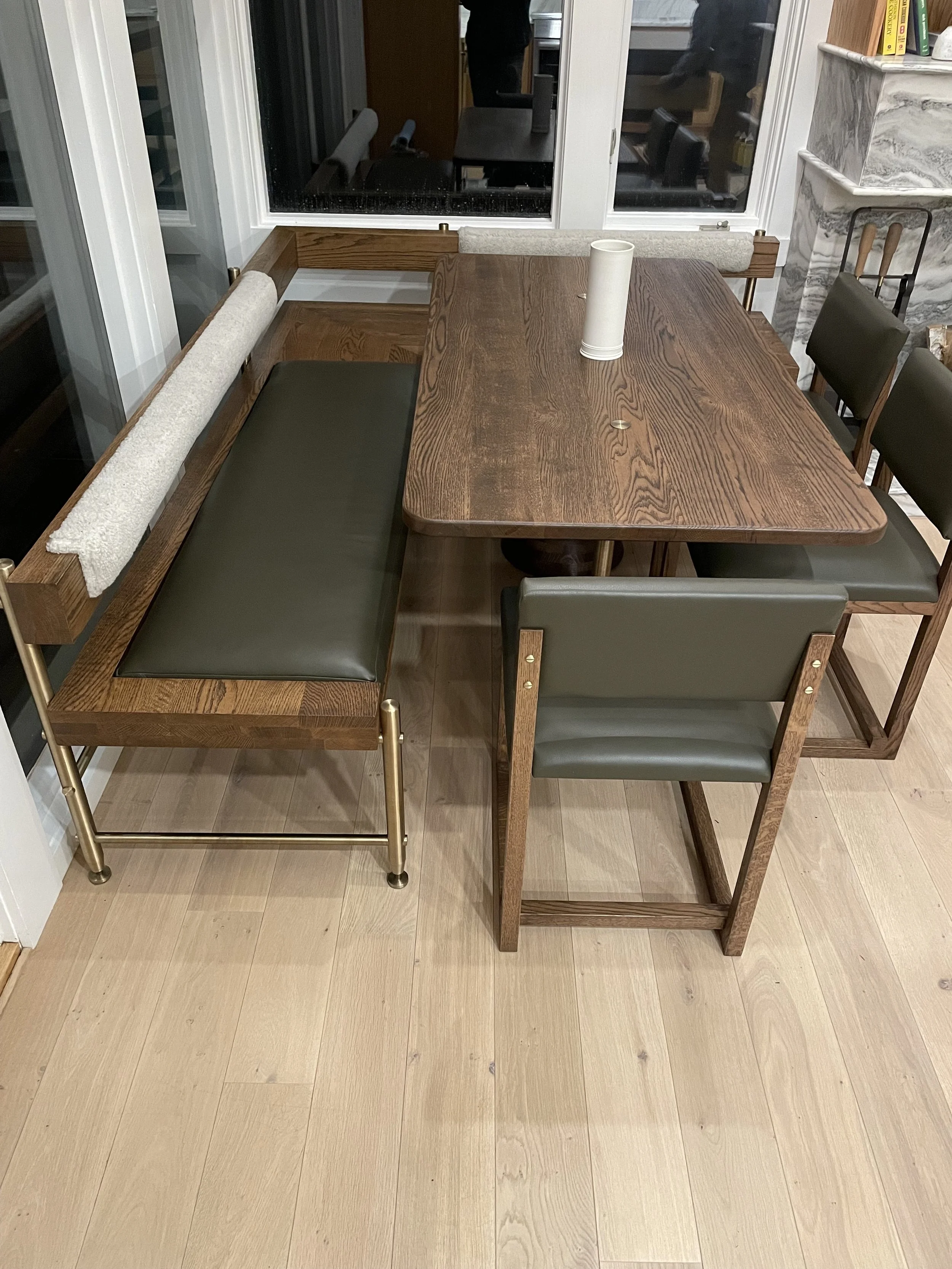 A wooden dining table with a roll of paper towels on top, surrounded by chairs and a bench, in a room with large windows and light wooden flooring.