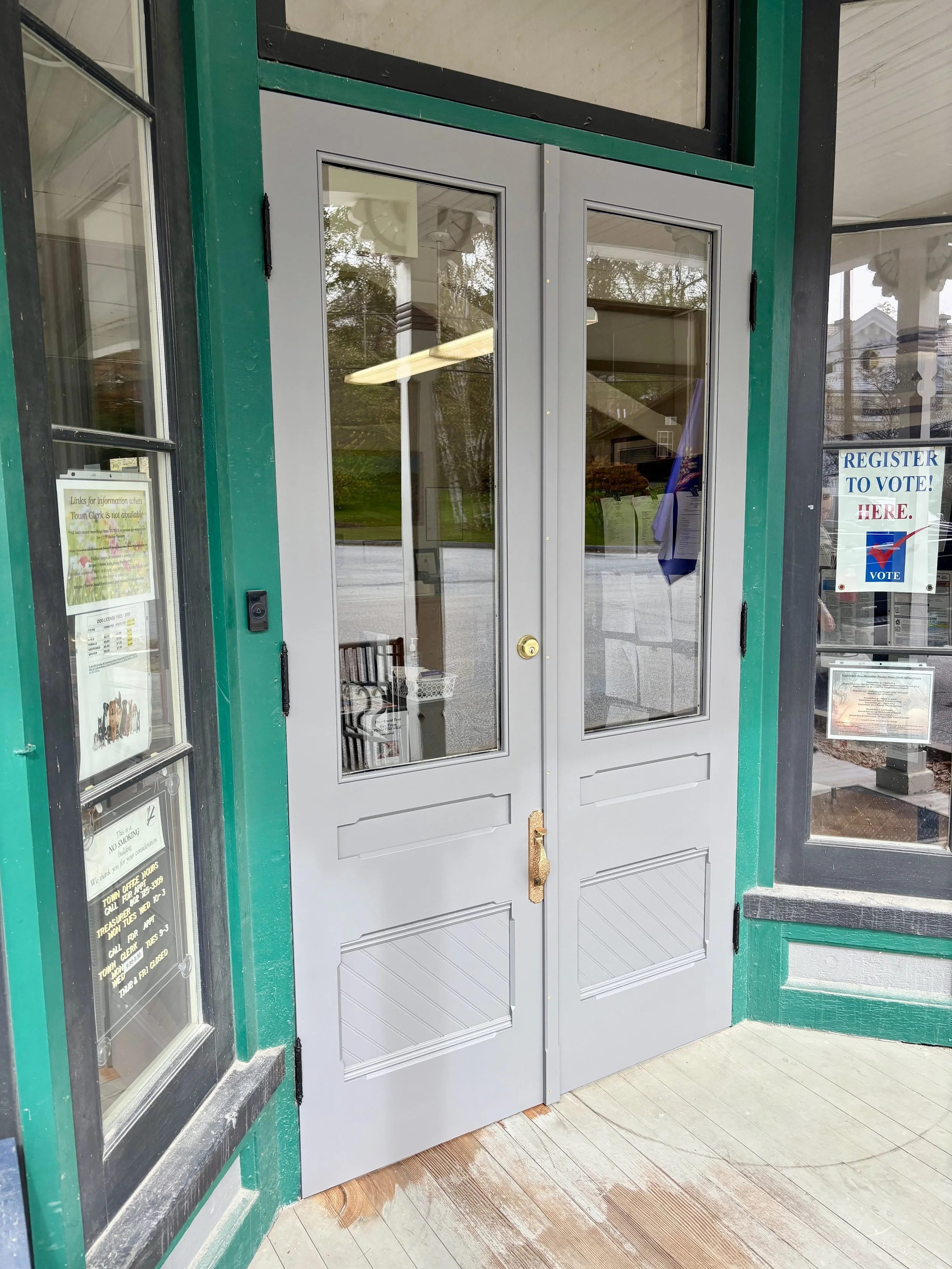 Exterior of a building with a closed gray double door with glass panes, set in a green and black frame, and a small wooden porch with a worn finish.
