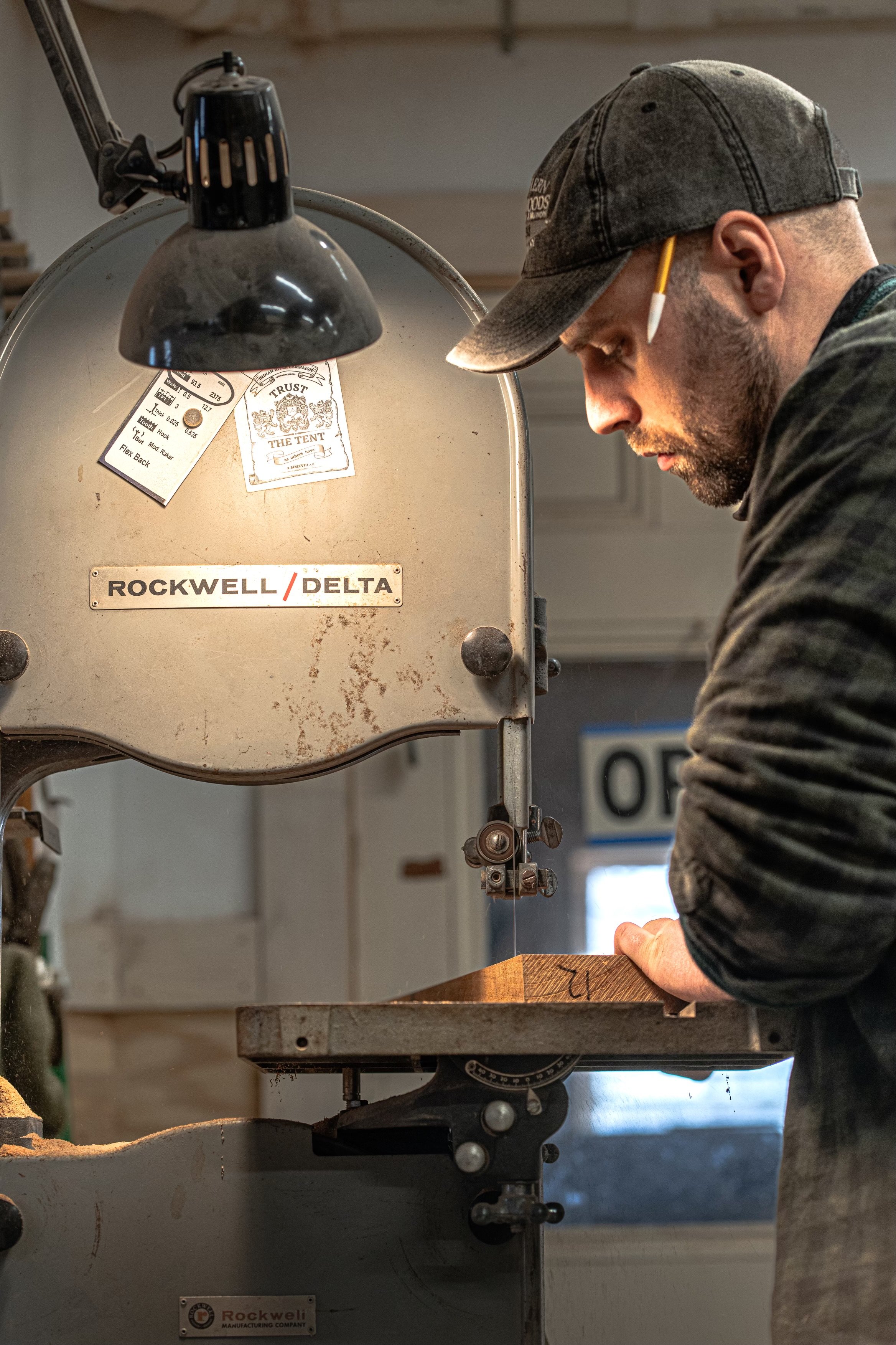 A man wearing a cap and long sleeve shirt operating a bandsaw to cut a piece of wood in a woodworking shop.