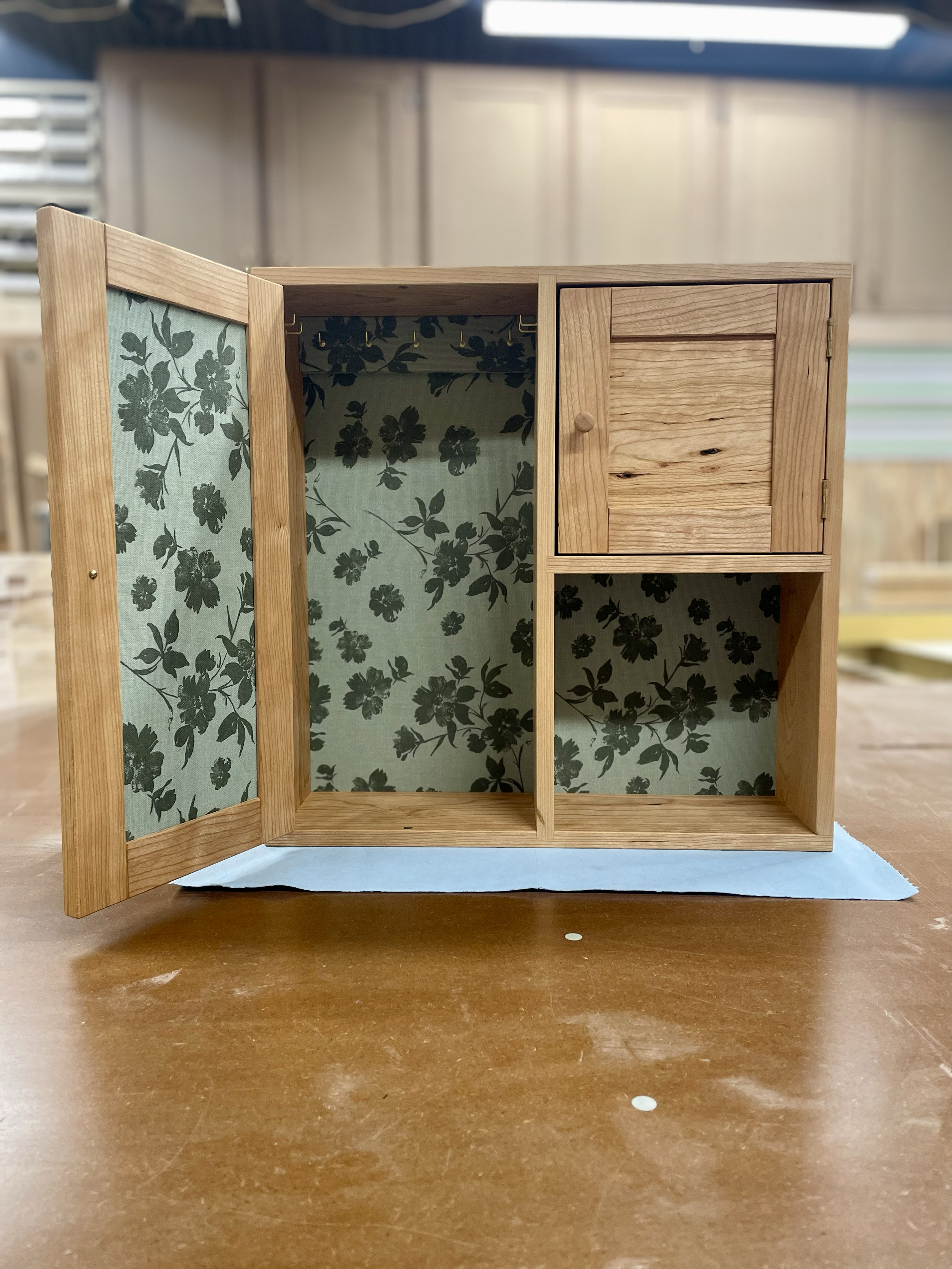 A small wooden cabinet with a floral patterned interior and a small door, placed on a wood table.