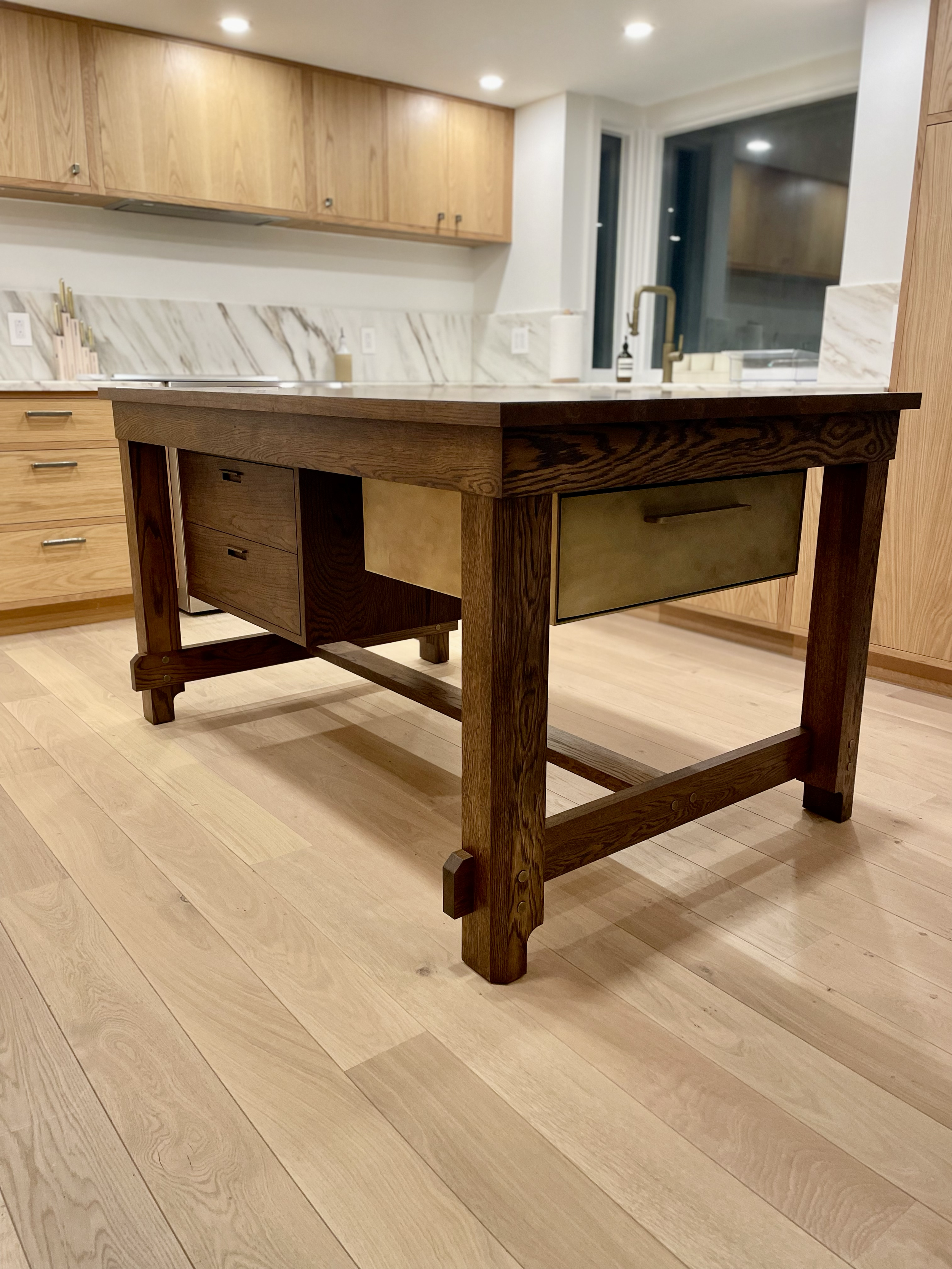 A wooden kitchen island with drawers and a glass-front cabinet, situated on a light wood floor in a modern kitchen with wooden cabinets and marble countertops.