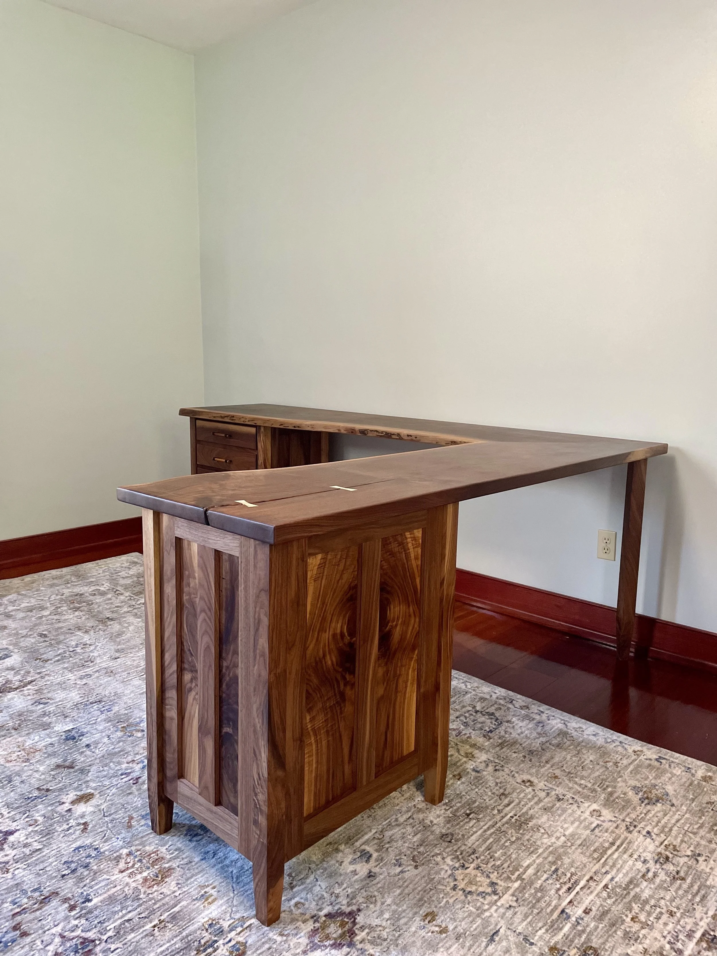 An L-shaped wooden desk with some drawers, set in a room with light-colored walls and a patterned rug on a dark hardwood floor.