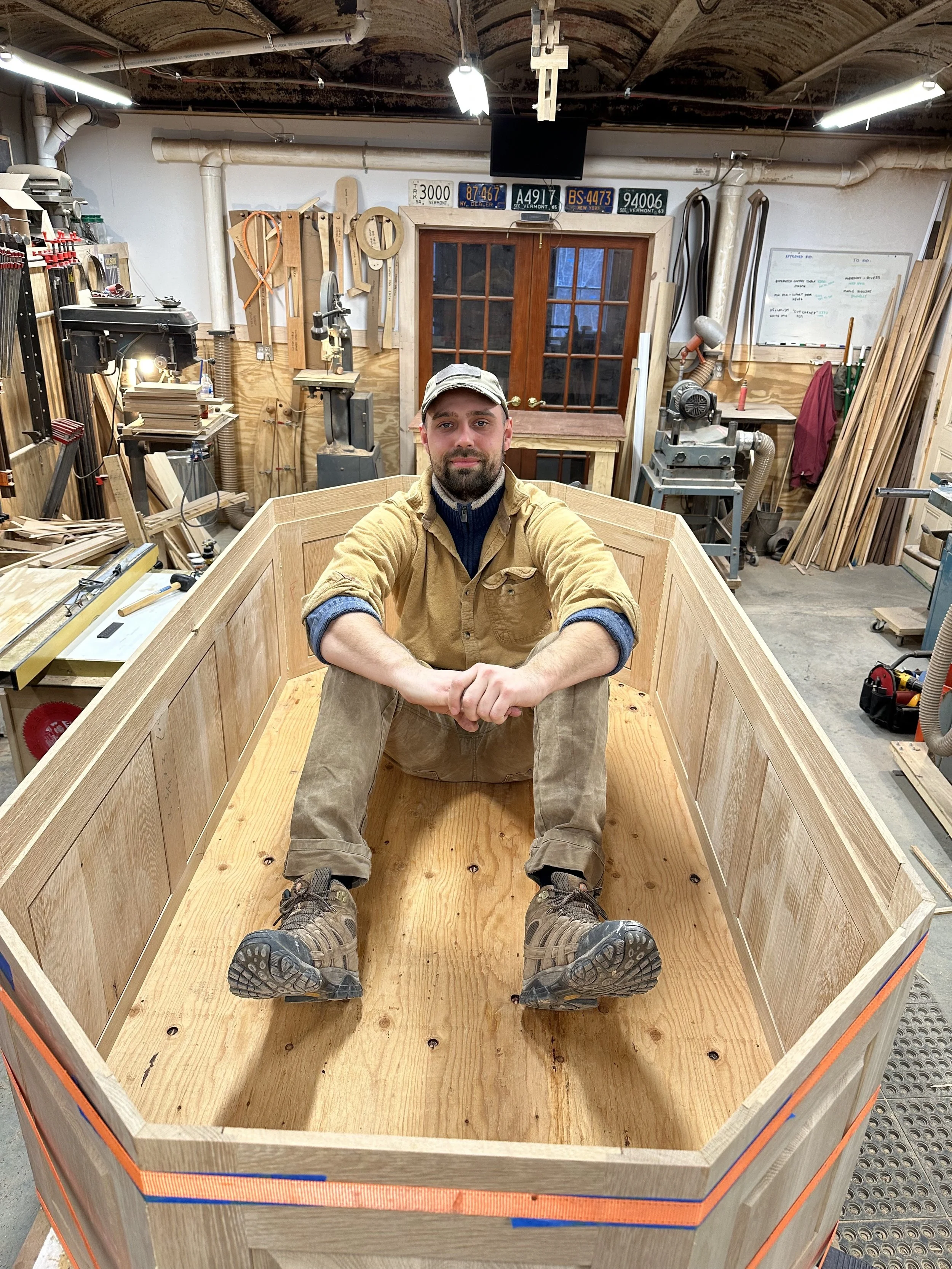 A man sits inside a large wooden boat frame in a woodworking shop. He wears a tan work shirt, khaki pants, sturdy work boots, and a cap, and is looking at the camera with a slight smile.