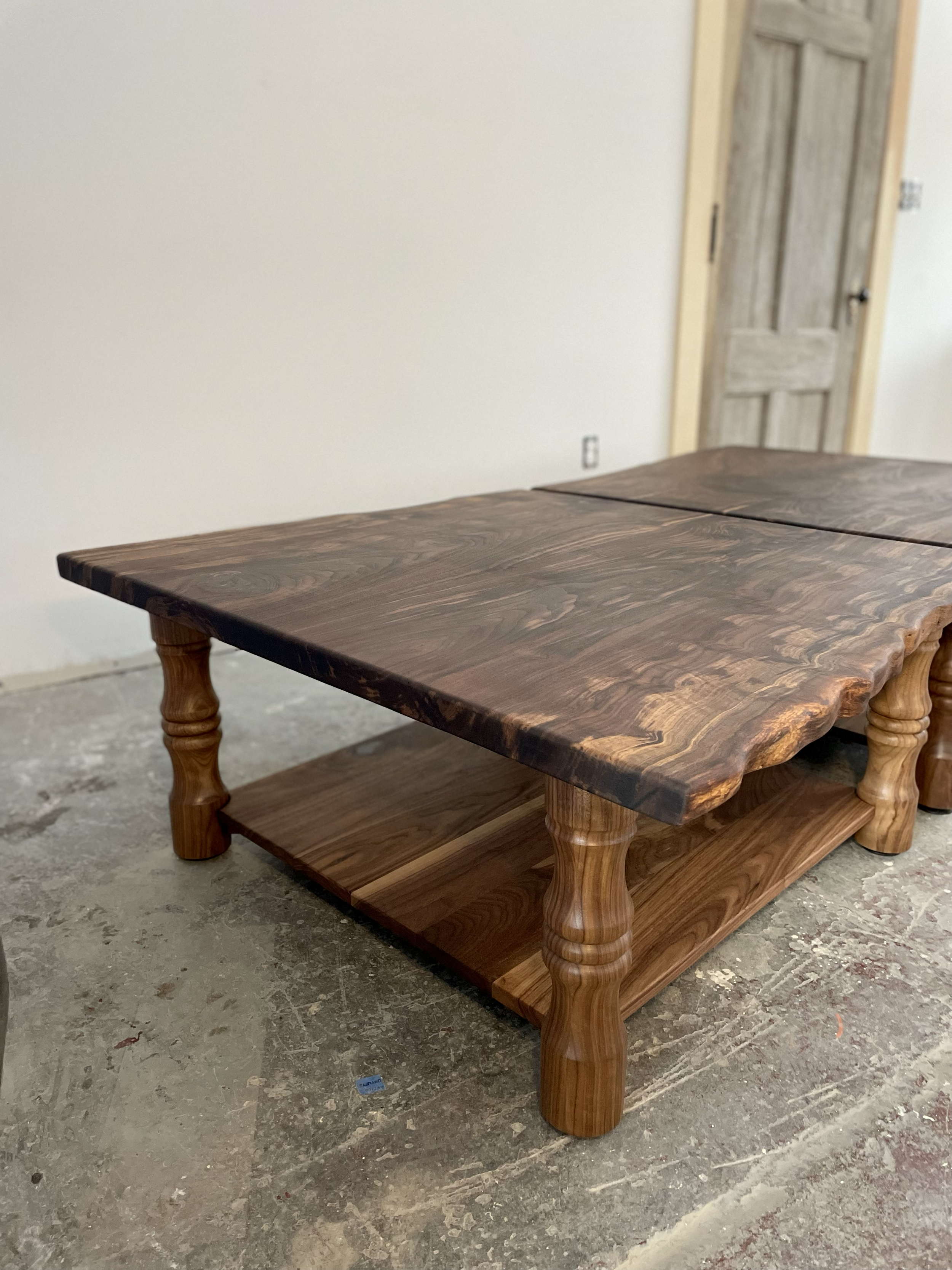 Wooden coffee table with a thick, polished tabletop and a lower shelf, situated on a concrete floor in an unfinished room with a wooden door in the background.