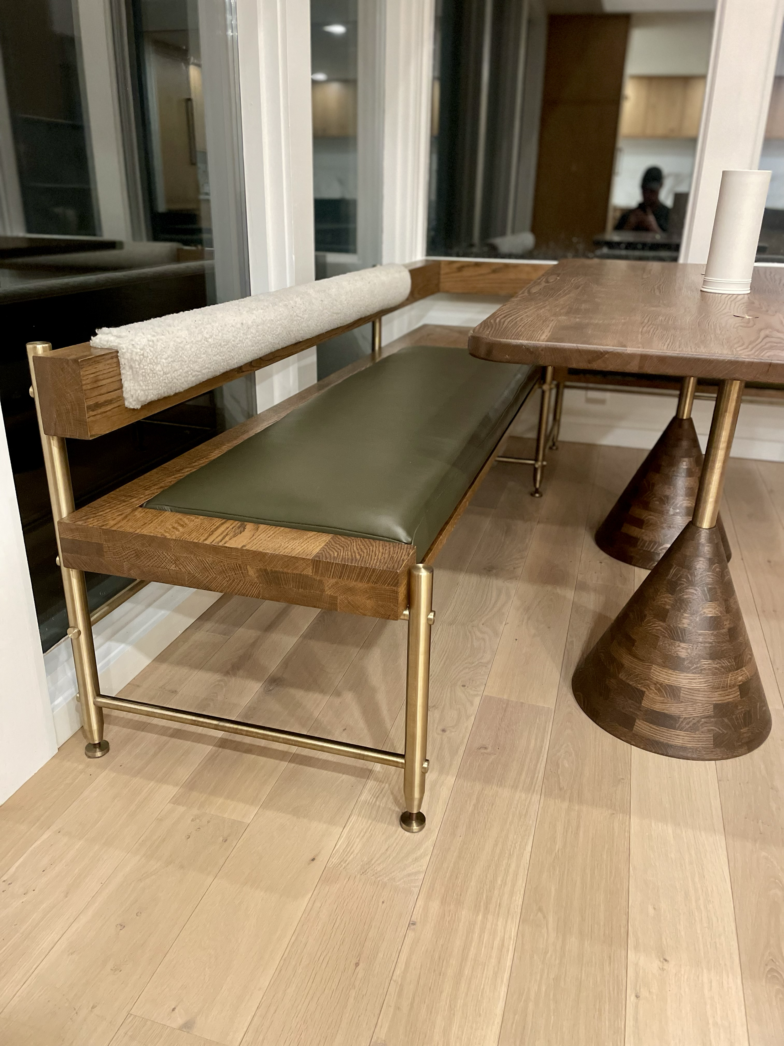 A wooden bench with a green cushion and a fabric-covered armrest in a room with light wood floors and a large window.