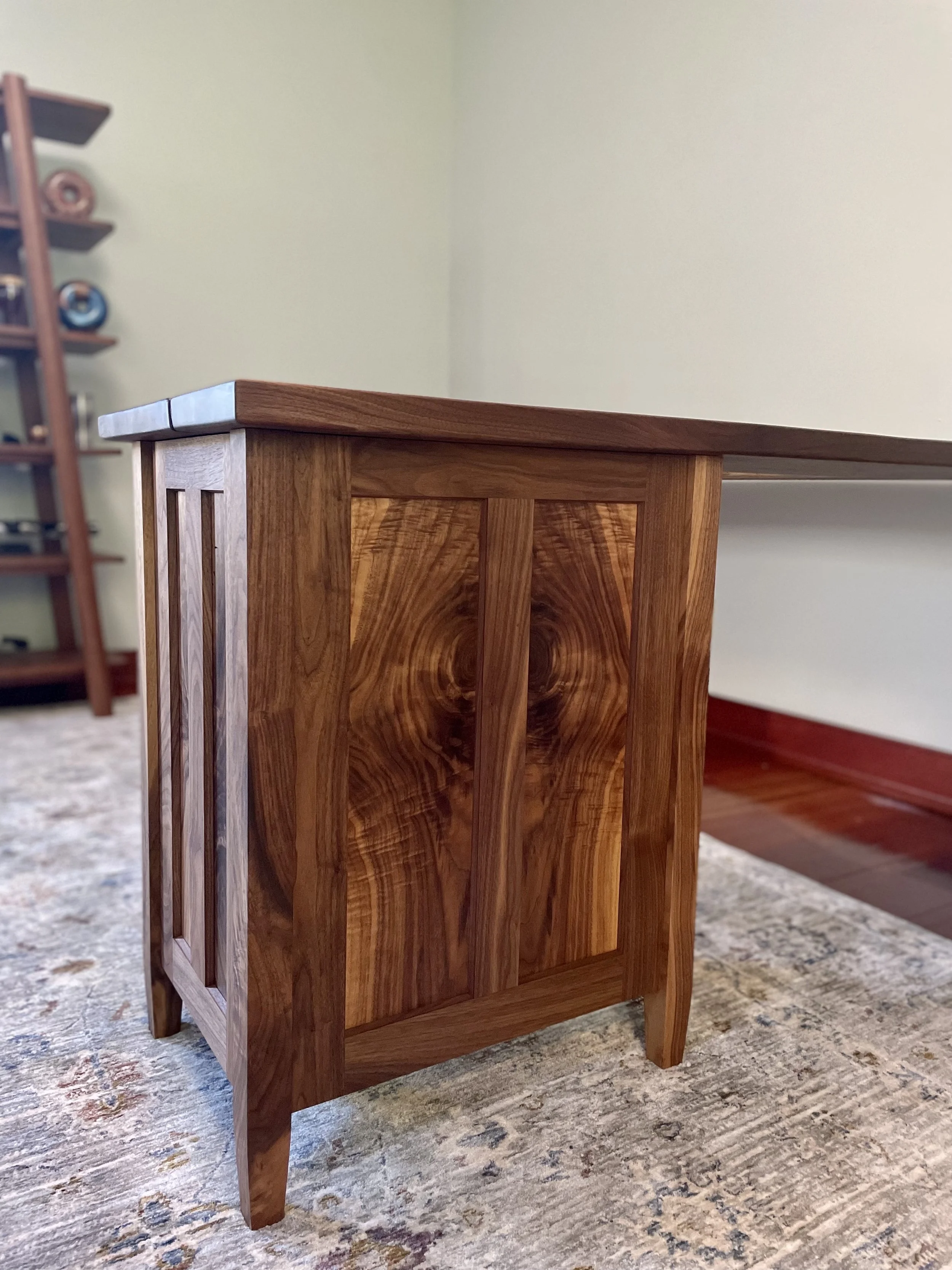 Close-up of a wooden side table with a smooth top and vertically paneled sides, placed on a textured rug in a room with a bookshelf in the background.