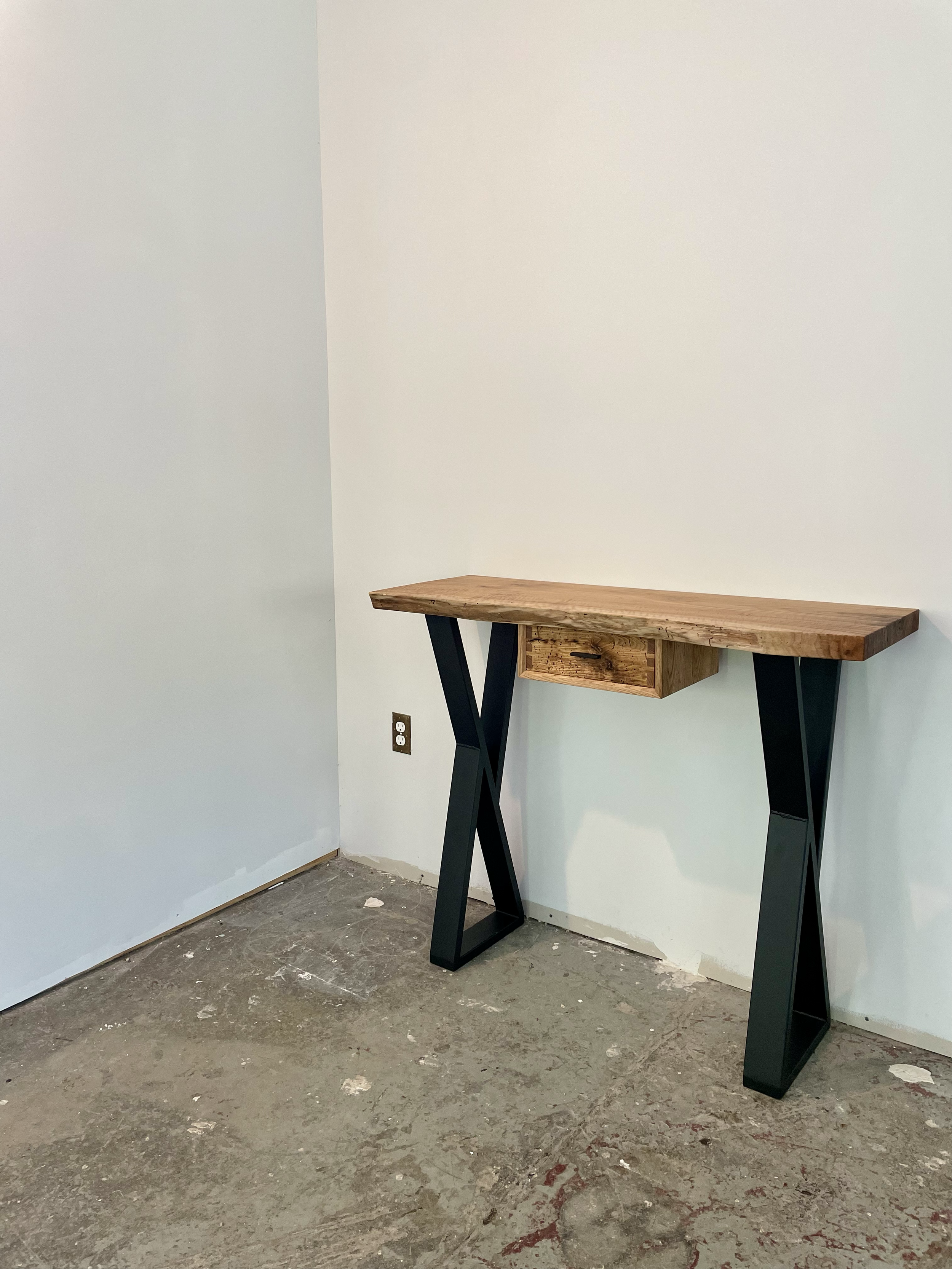 A small wooden desk with black metal legs and a single small drawer, positioned against a white wall in a room with a concrete floor.