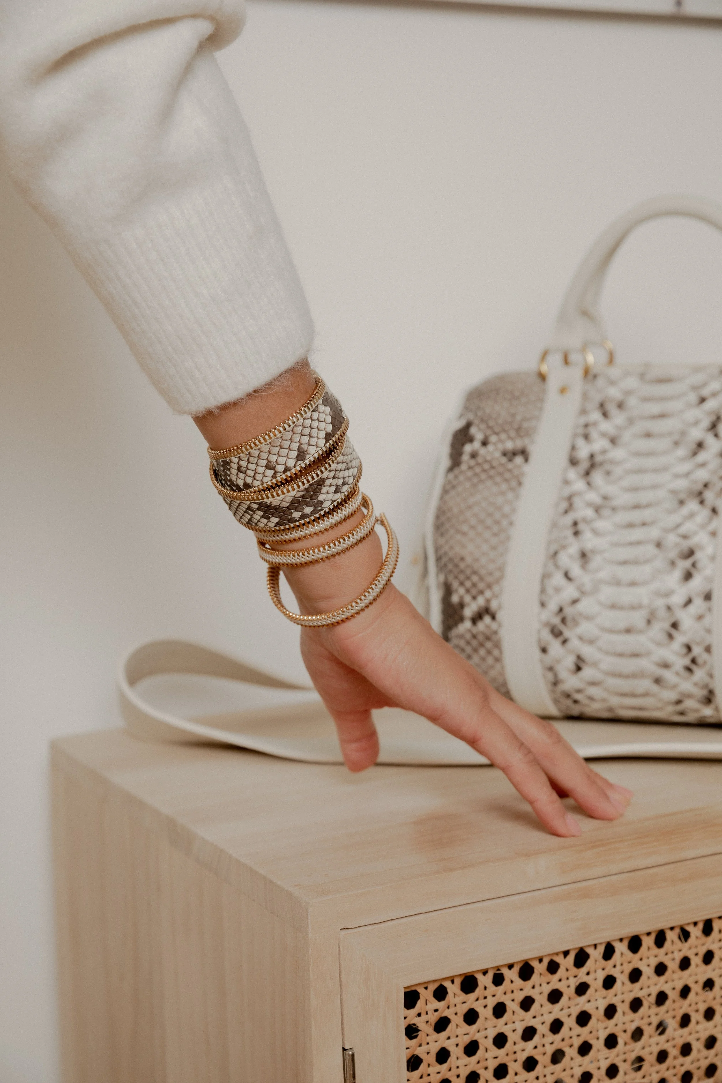 Une main féminine portant plusieurs bracelets dorés et à motif serpent, posée sur un meuble en bois avec une valise à motif serpent à côté d'une sac en bandoulière en raphia beige.