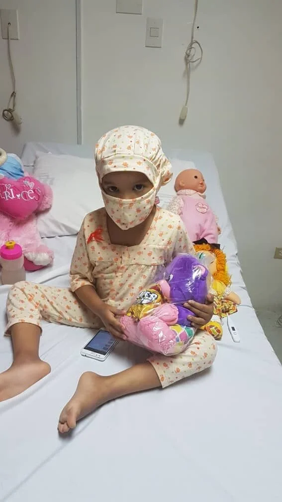 Niña en cama del hospital con mascarilla, usando piyama de flores, rodeada de muñecos y juguetes, sosteniendo una bolsa de dulces y con un teléfono móvil a su lado.