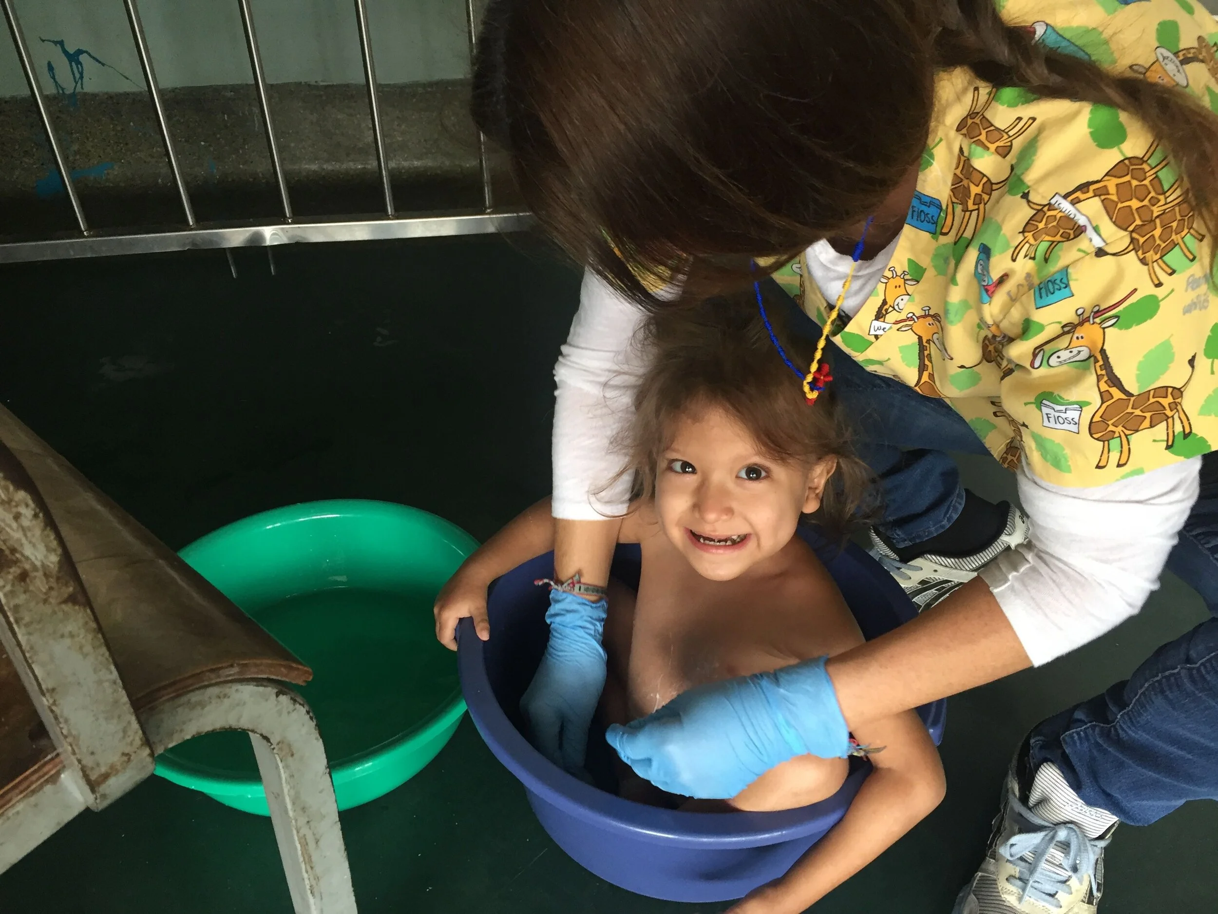 Dos niños y una adultez en un ambiente hospitalario, con una tina de plástico y un recipiente grande, probablemente en un procedimiento de lavado o cuidado infantil.