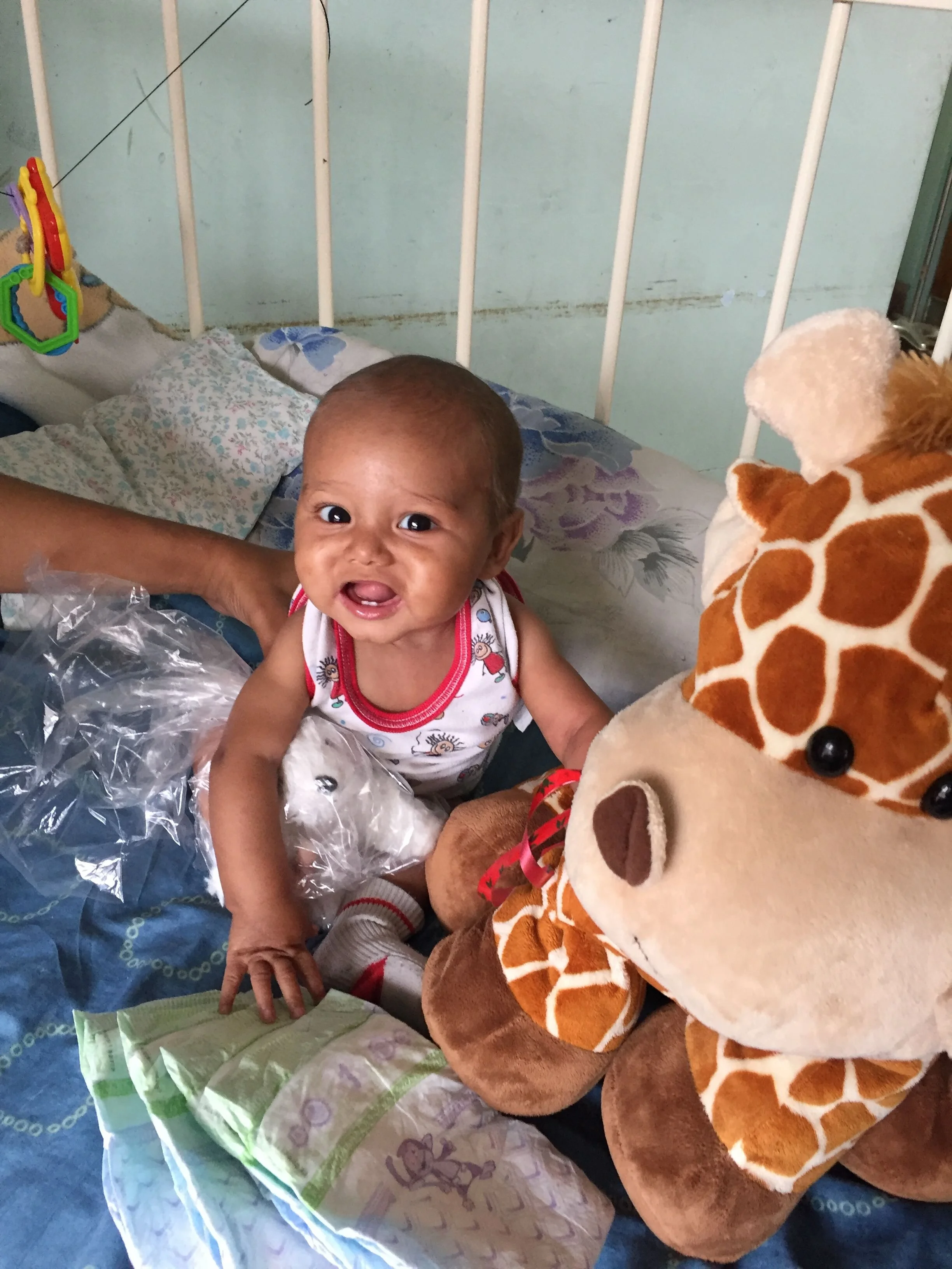 Un bebé sonriente en la cama rodeado de peluches y una manta. 