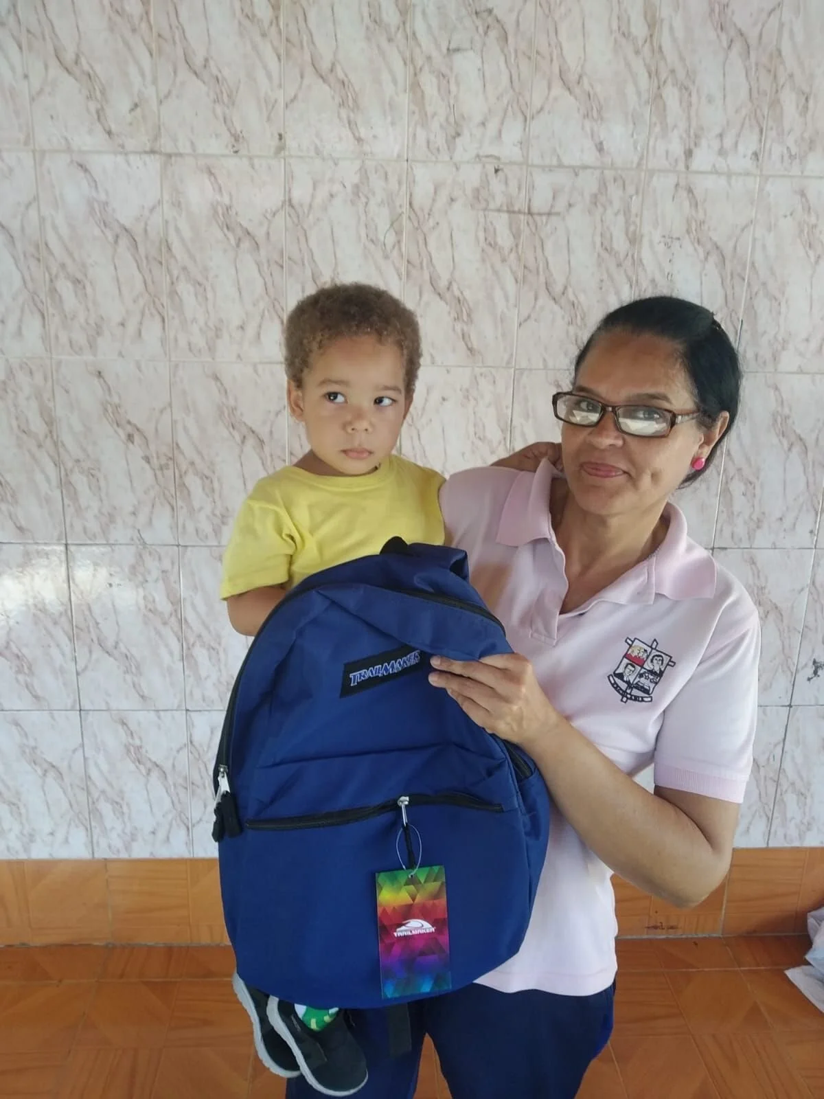 Una mujer y un niño posan juntos, la mujer sostiene una mochila azul y el niño lleva una camiseta amarilla con pantalones deportivos negros con franjas blancas. La mujer usa gafas, una blusa rosa y pantalones oscuros, y está junto a una pared de azul