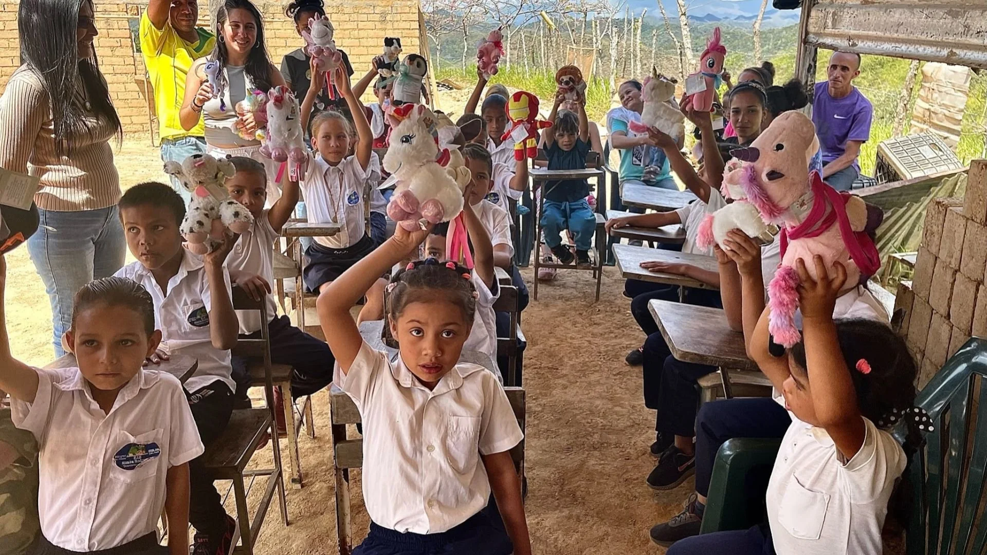 Niños en una clase rural sosteniendo peluches y muñecos, algunos adultos observando, en un ambiente al aire libre y rústico.
