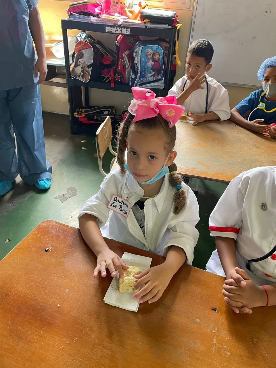 Niña con un lazo grande rosa en el cabello, sentada en una mesa en una escuela, con una etiqueta que dice ‘Doctora Zae Perla’, sosteniendo un trozo de pastel. En el fondo, otros niños y una persona con ropa de protección están en un aula con mochilas