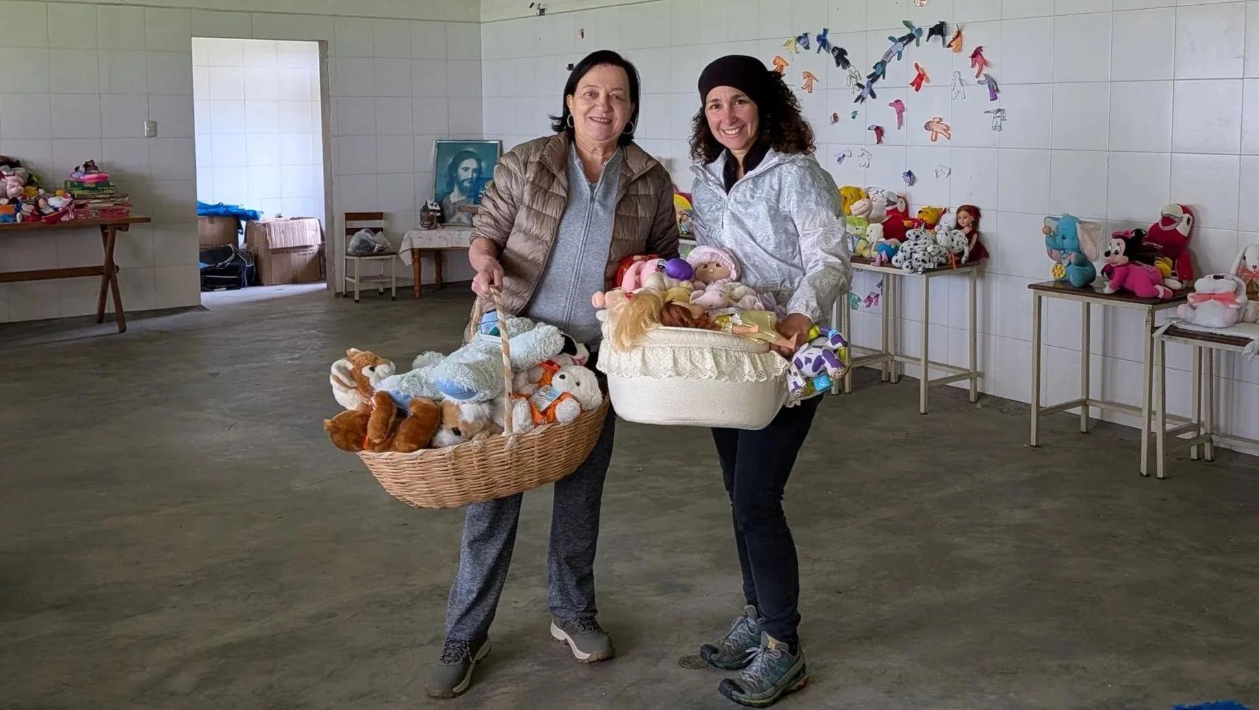 Dos mujeres sosteniendo cestas llenas de muñecos de peluche en un salón con paredes de azulejo y mesas con más muñecos fuera de vista y en las esquinas, en un evento o mercadillo de juguetes.