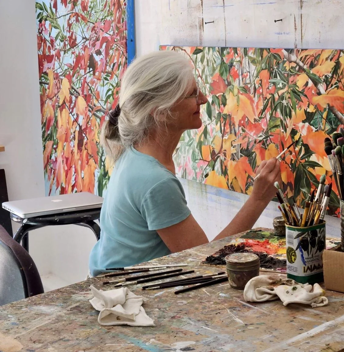 Artist Madeleine Wood, wearing glasses and a light blue shirt, painting a large canvas of colorful orange and yellow leaves with green leaves and branches in the background in an art studio.