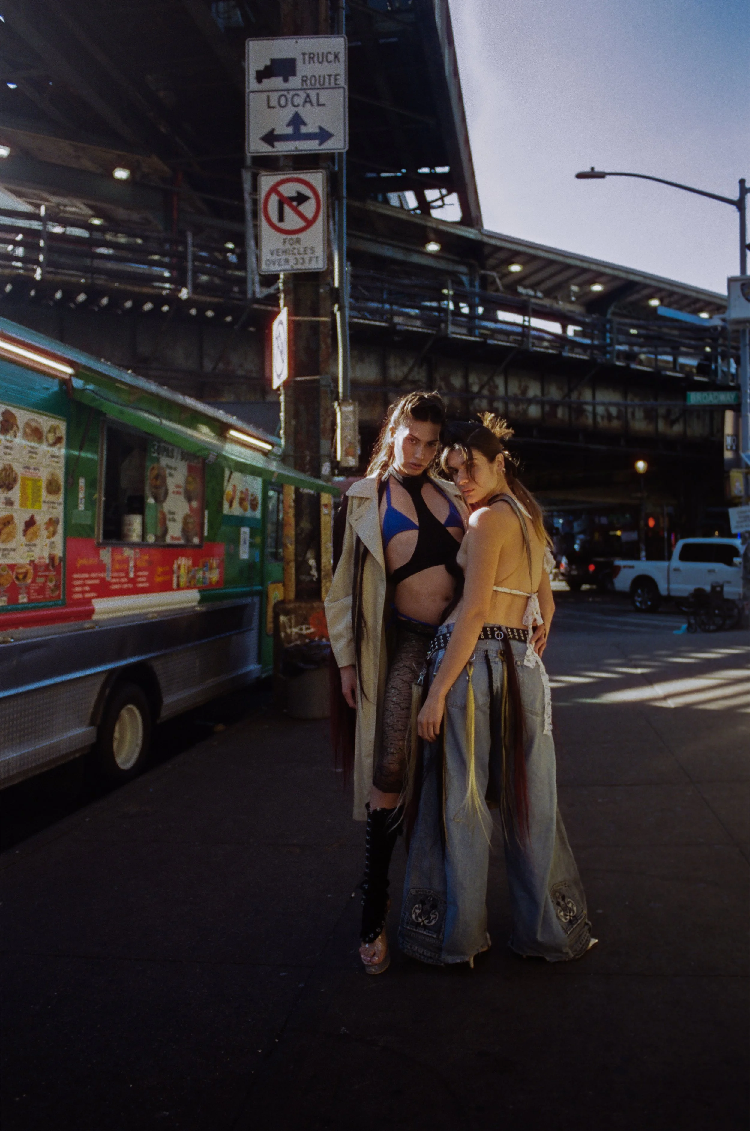 Two young women with dark hair posing on a city street, dressed in edgy, revealing clothing, with a food truck and street signs in the background during evening or dawn.