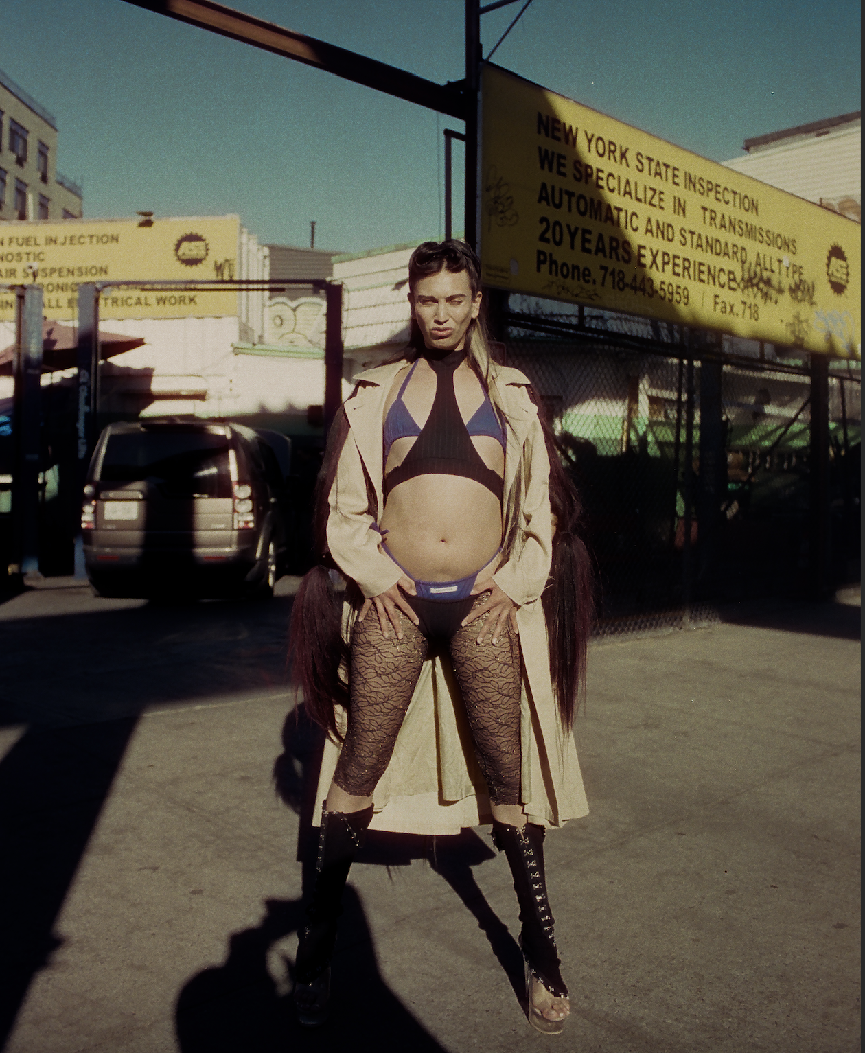 A woman standing outdoors in a dramatic pose, wearing unconventional clothing including fishnet stockings, a crop top, and a trench coat, with a cityscape and signs in the background.