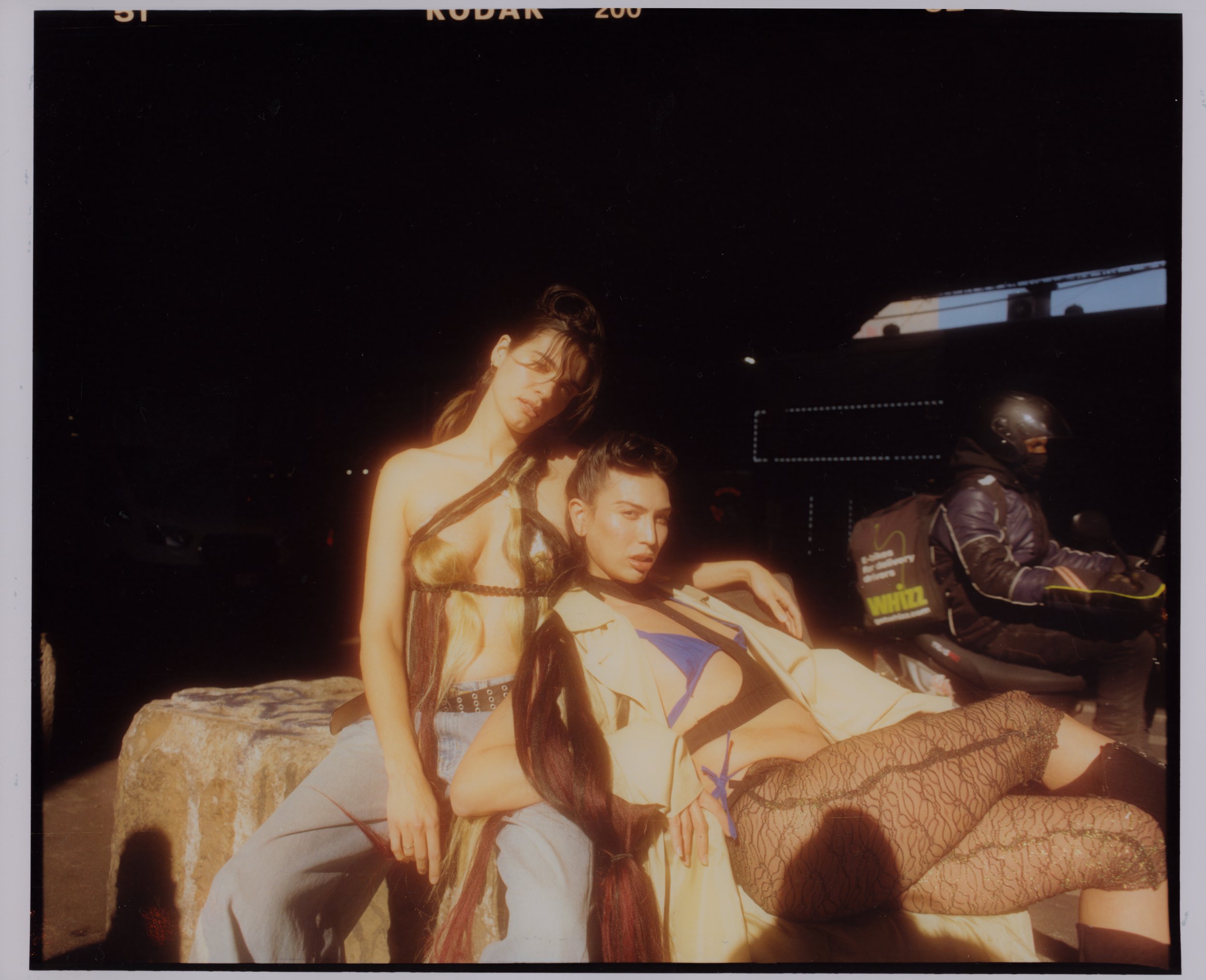 Three models posing outdoors near a large rock, with a dark background and a structure with lights in the distance. The models are dressed in fashion outfits, including fishnet stockings and colorful clothing.
