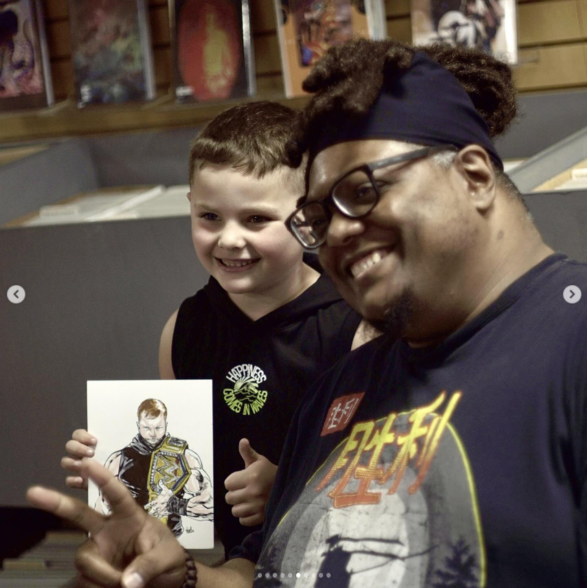 A young boy and an adult man smiling and posing for a photo. The boy is holding a drawing of a wrestler with a championship belt, and the man is making a peace sign. They are in a room with books or artwork on shelves in the background.