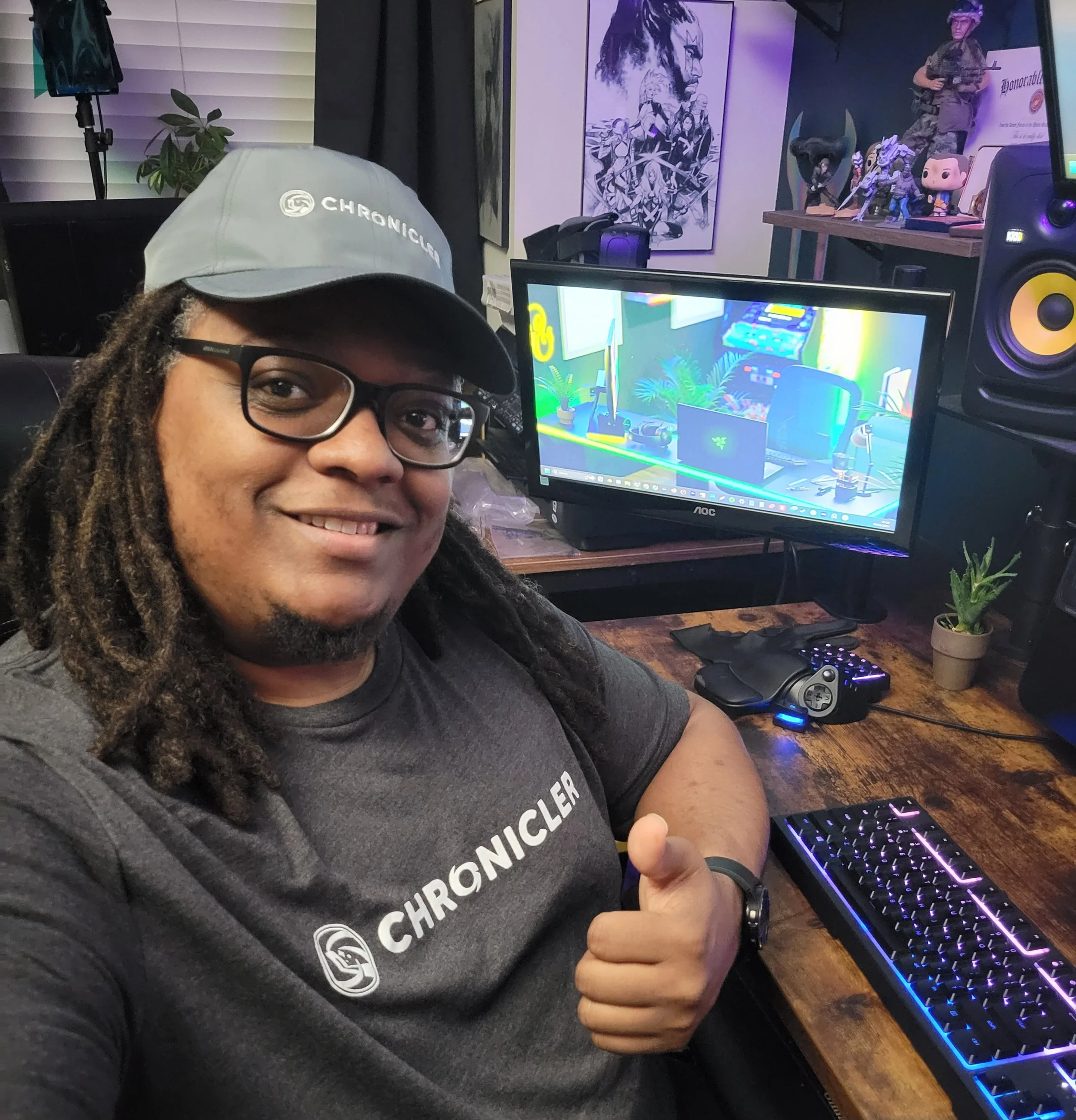A person wearing glasses and a gray cap with a Chronology logo, sitting at a desk with a computer, giving a thumbs-up. In the background, there are monitors, speaker, and various collectibles including action figures, artwork, and a small plant.