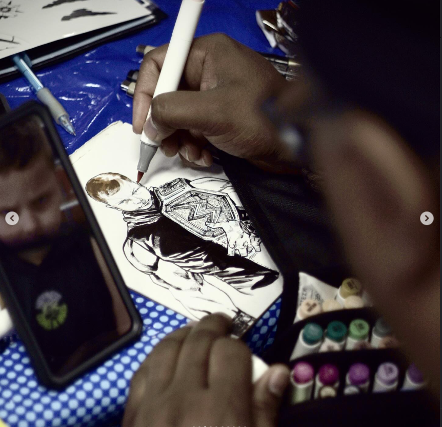Artist drawing a black and white illustration of a male comic book character with a high-collared jacket and armor on a sketchpad, with art supplies and a smartphone nearby.