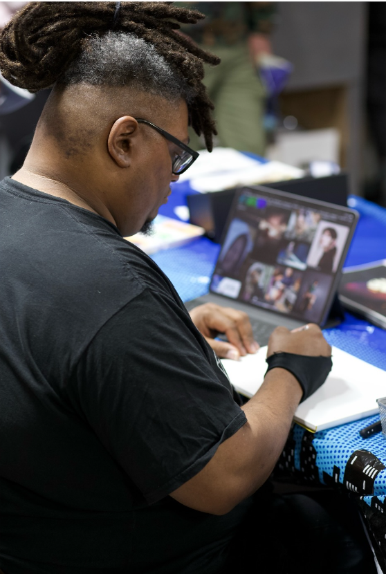A person with dreadlocks and glasses writing in a notebook at a table with a laptop displaying a collage of images.