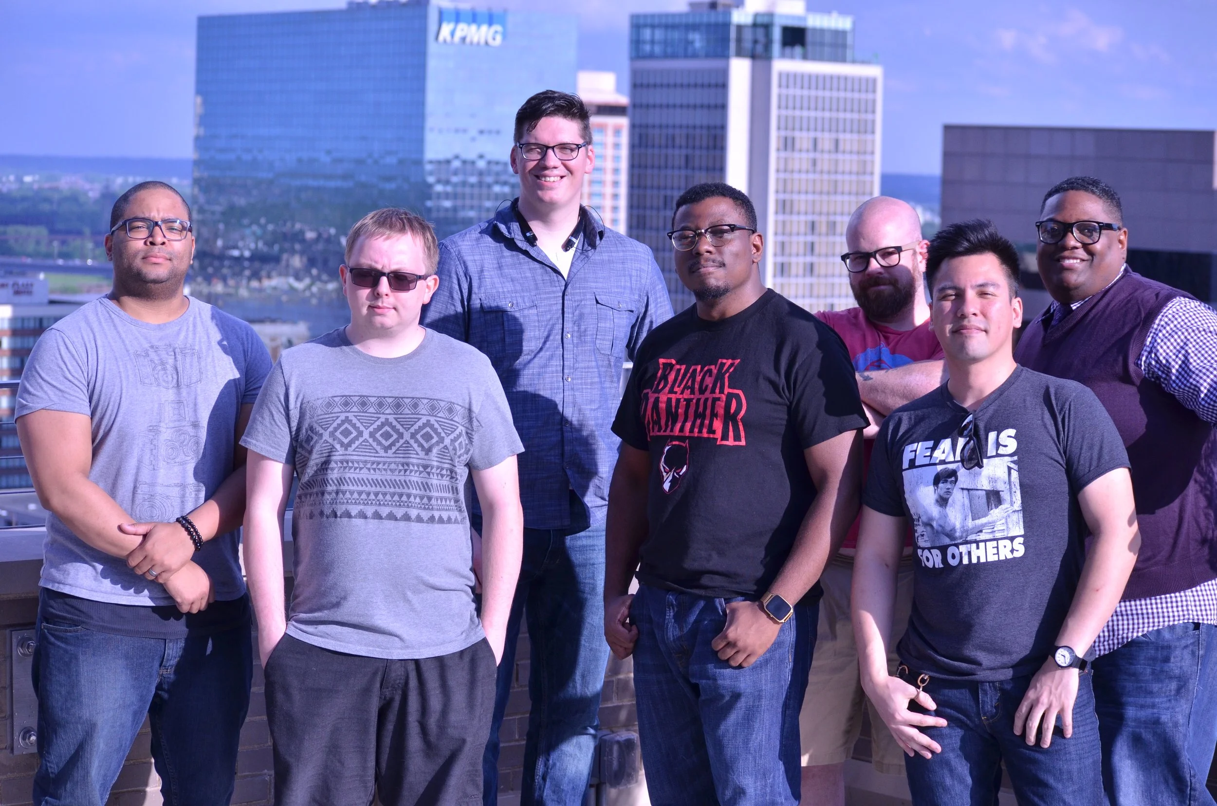Group of eight diverse men standing on a city rooftop with tall buildings in the background, smiling and posing for the camera.