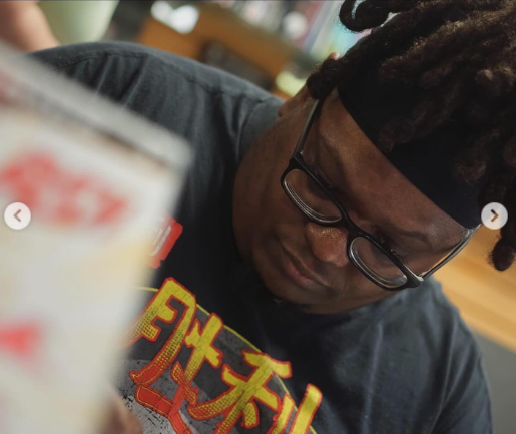 A young man with dreadlocks, glasses, and a headband looking down at his food in a casual indoor setting.