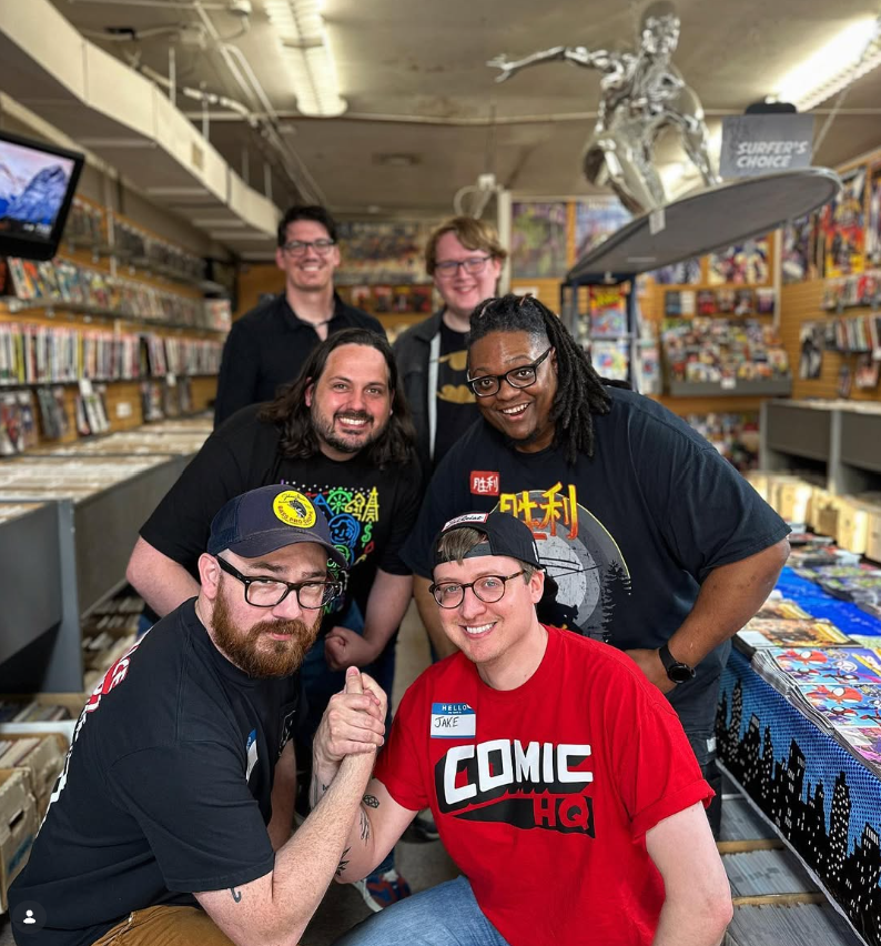 Group of six people posing together in a comic book store, with comic books and artwork displayed on shelves around them, and a futuristic silver robot figure hanging from the ceiling.