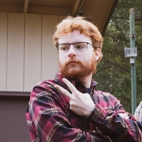 Young man with red hair and beard wearing glasses and a plaid shirt, making a peace sign with his fingers.
