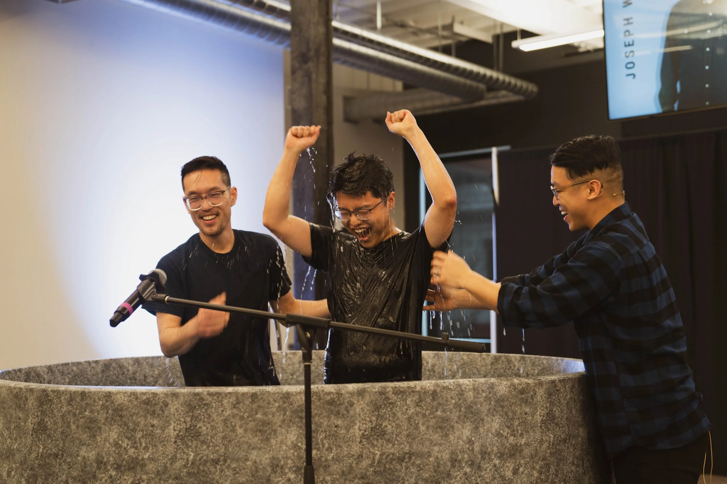 Three young men are celebrating at a baptism ceremony in a church or community center, with one person being baptized in a water basin as water splashes around.