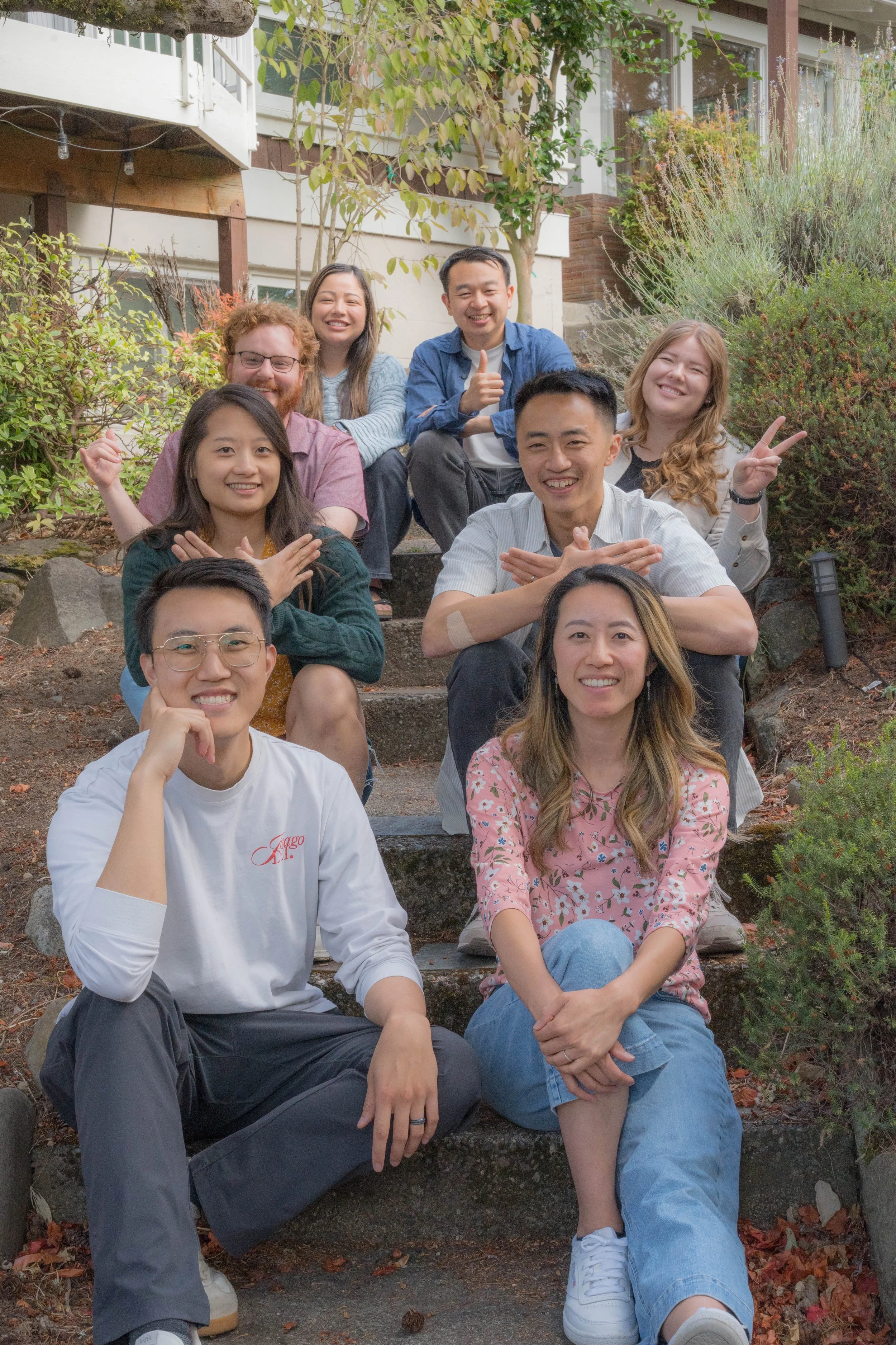 A group of diverse young adults sitting on outdoor steps in front of a house, smiling and making playful gestures, surrounded by bushes and trees.