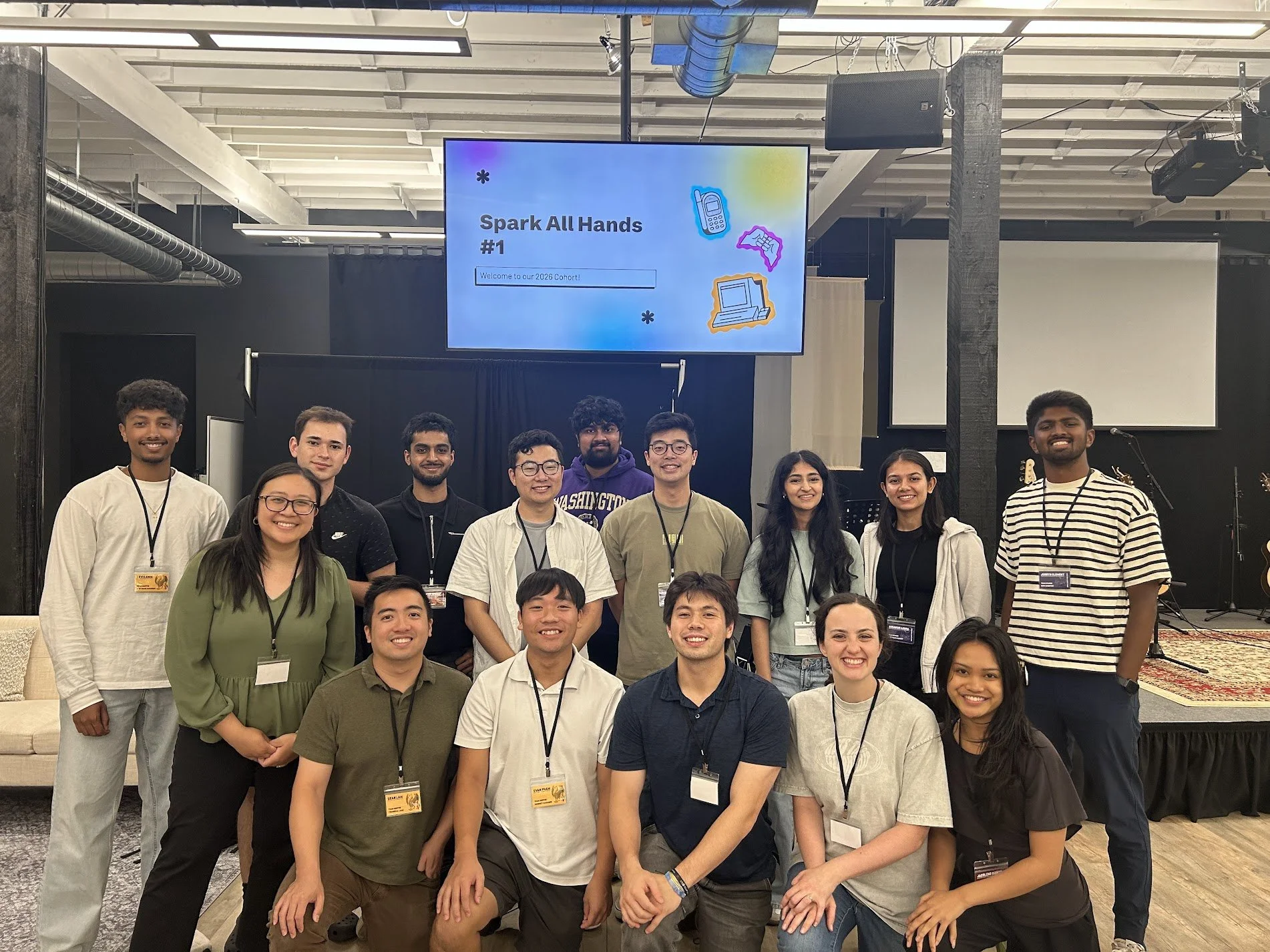 A group of young adults standing on a stage in a conference setting, smiling for a group photo. Behind them is a large screen displaying the text 'Spark All Hands #1' and an introduction message for a 2026 cohort, with colorful icons of a phone, a game controller, and a computer.