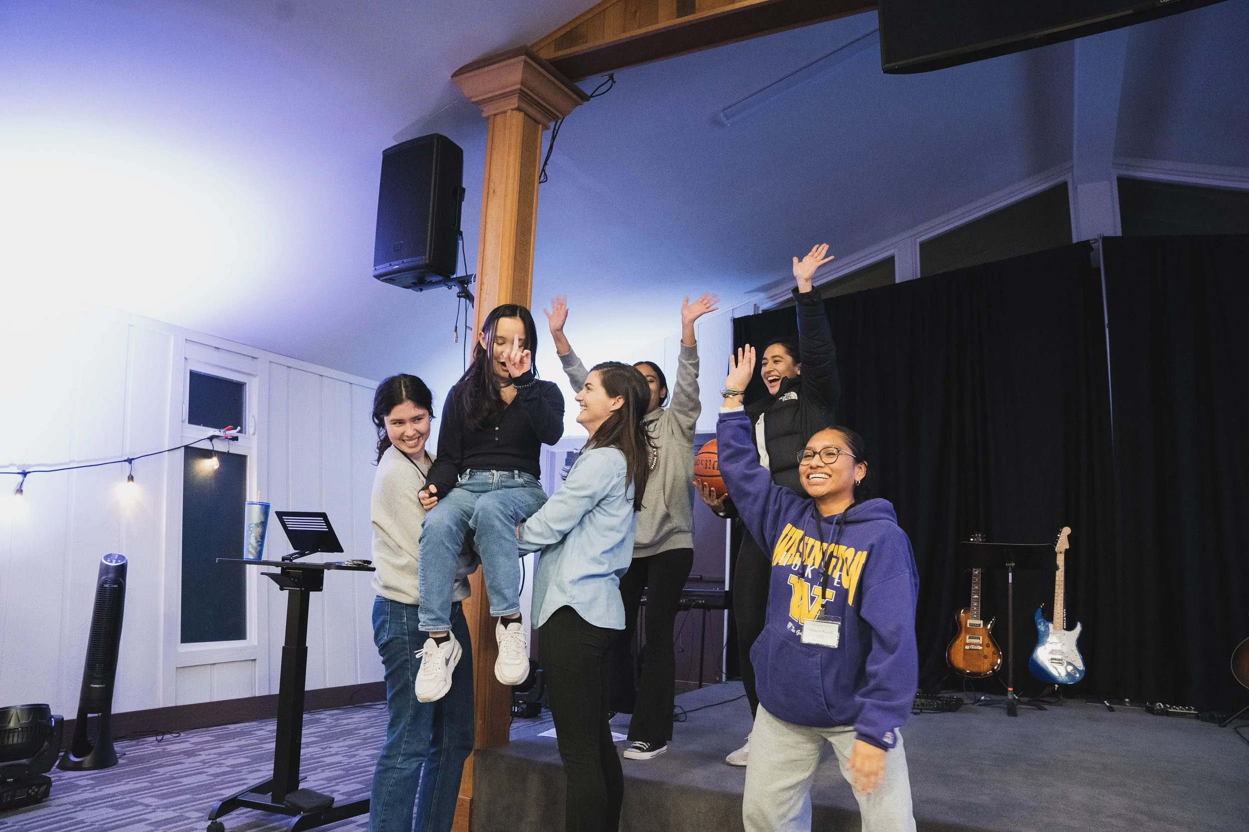 Group of young women joyfully celebrating indoors, with some raising hands and one being held up by others, in a room with musical instruments on stage in the background.