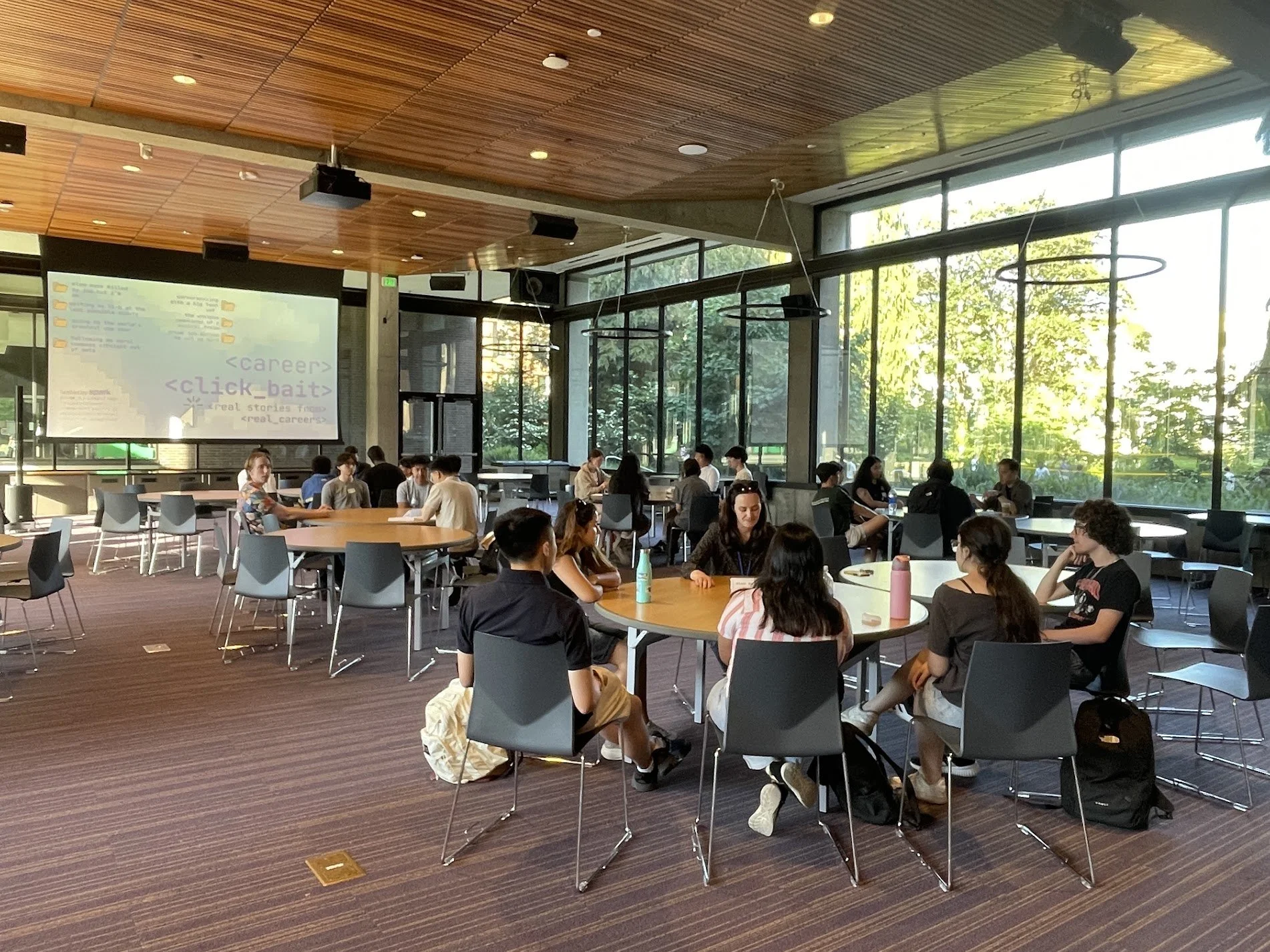 A large group of people sitting around tables in a modern, well-lit indoor space with large windows showing greenery outside, and a presentation projected on a screen.