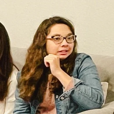 Woman with long curly hair and glasses sitting on a couch.