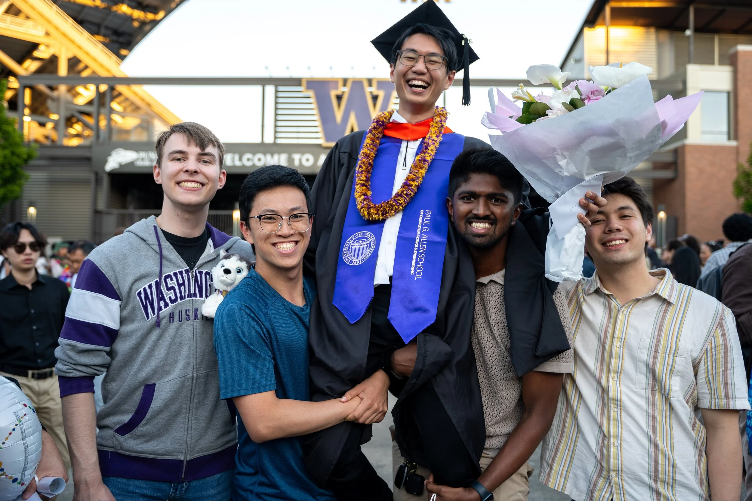 Group of five diverse young adult guys celebrating a graduation outside, with one wearing a graduation cap and gown, holding a flower bouquet, and all smiling.