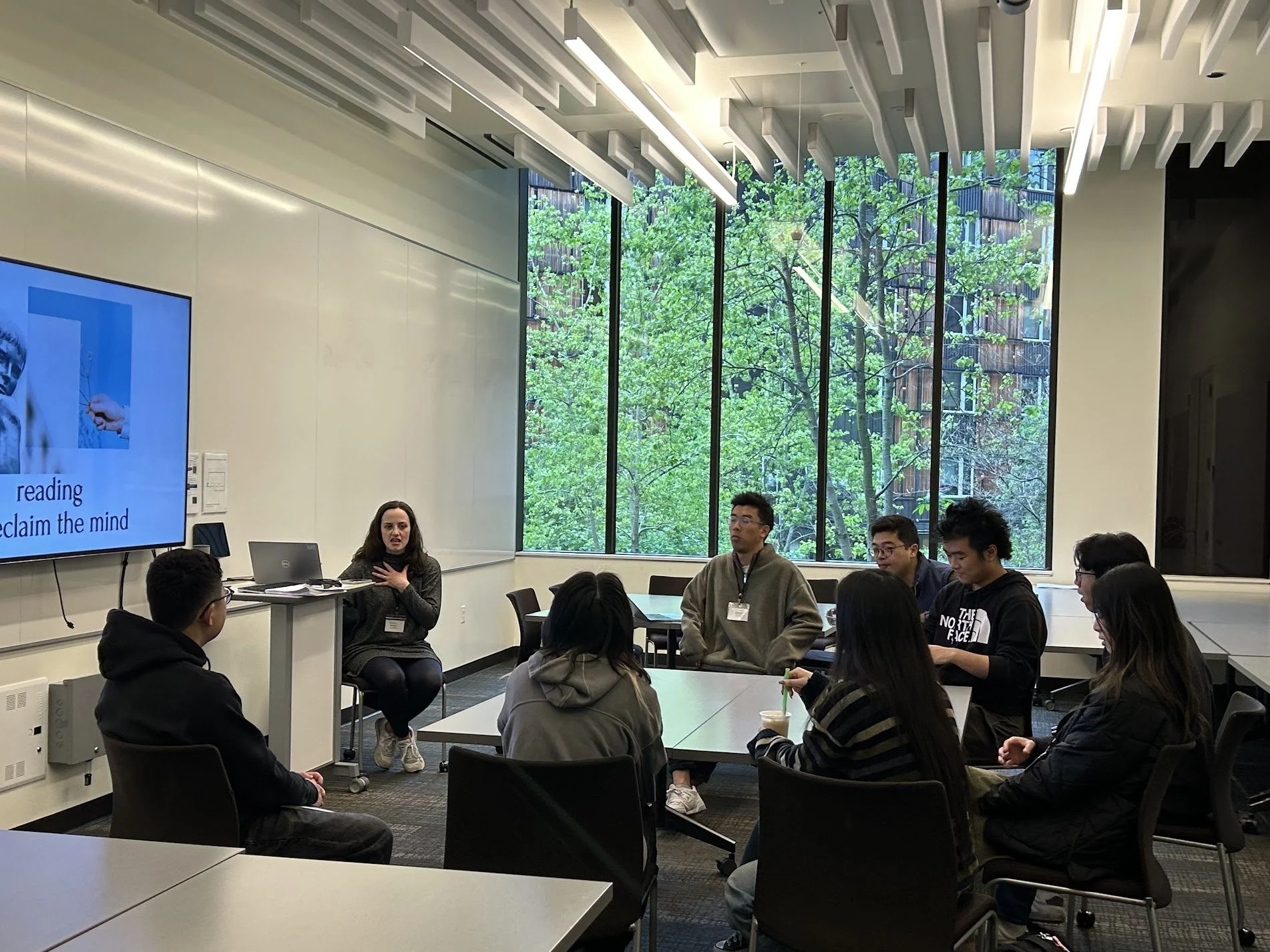 A group of diverse people participating in a discussion or workshop in a modern conference room with large window showing green trees outside. A woman is speaking while others listen attentively.