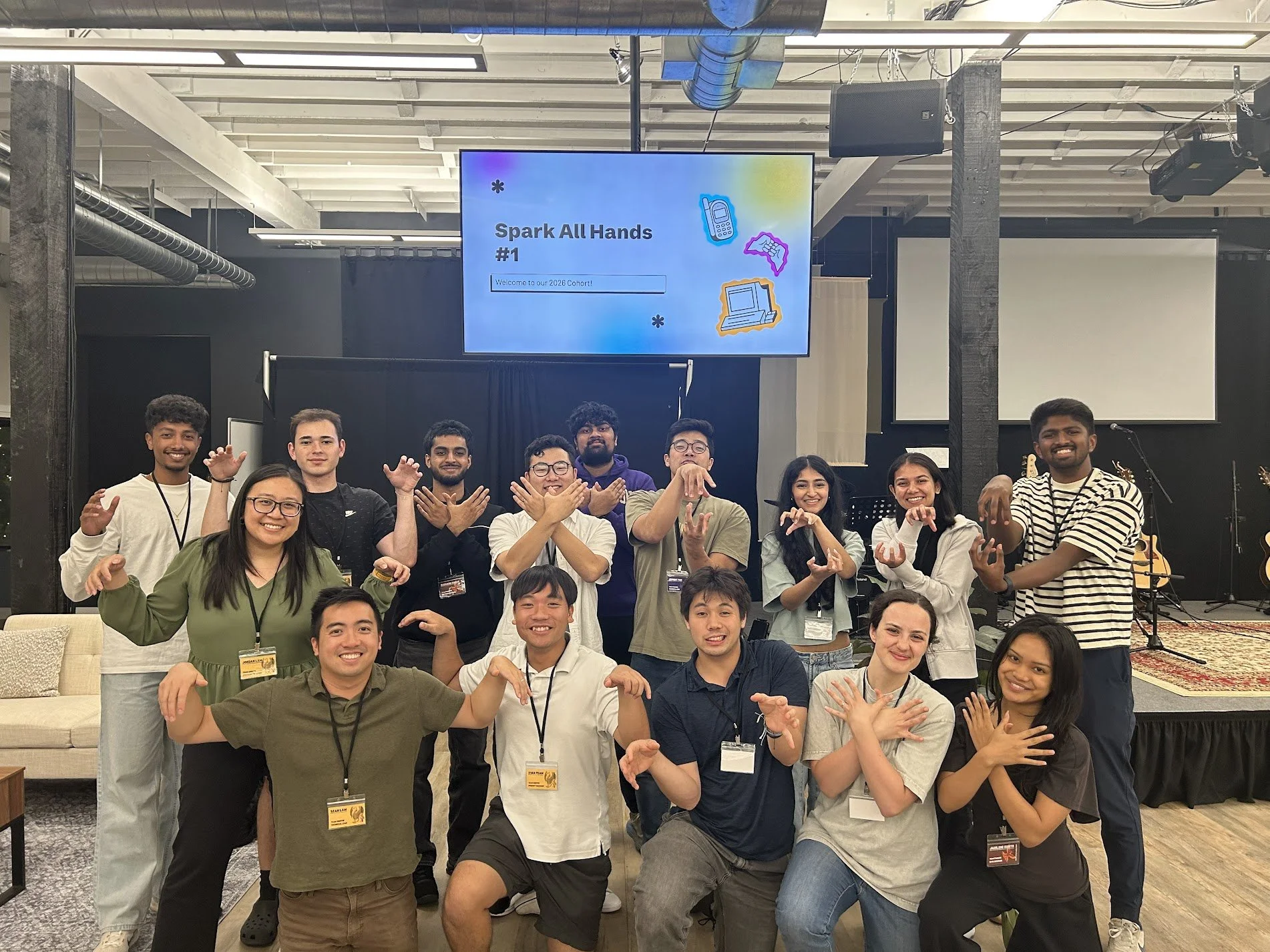 A diverse group of young adults gathered indoors at a conference or workshop, standing in front of a stage with a screen that displays a welcome message and graphics of electronic devices. They are smiling, making gestures, and wearing name badges.