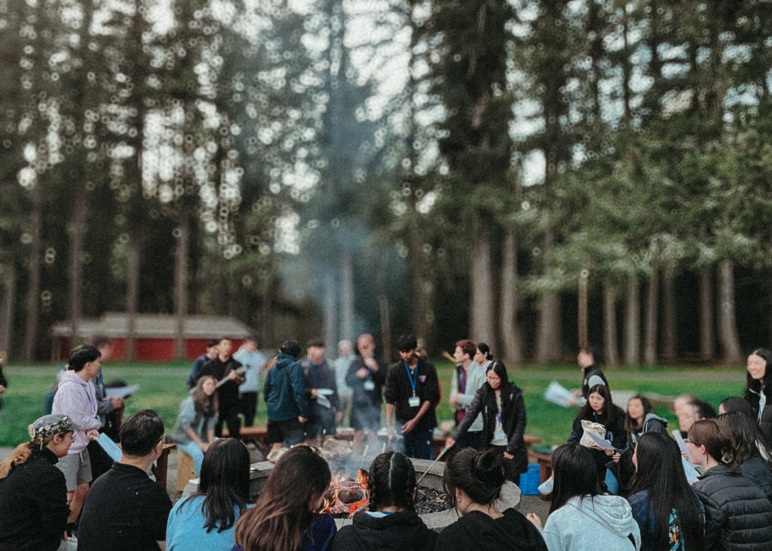 A group of people gathered outdoors around a campfire in a wooded area, some holding papers, with some standing and others sitting on benches, during a social or educational event in the evening.