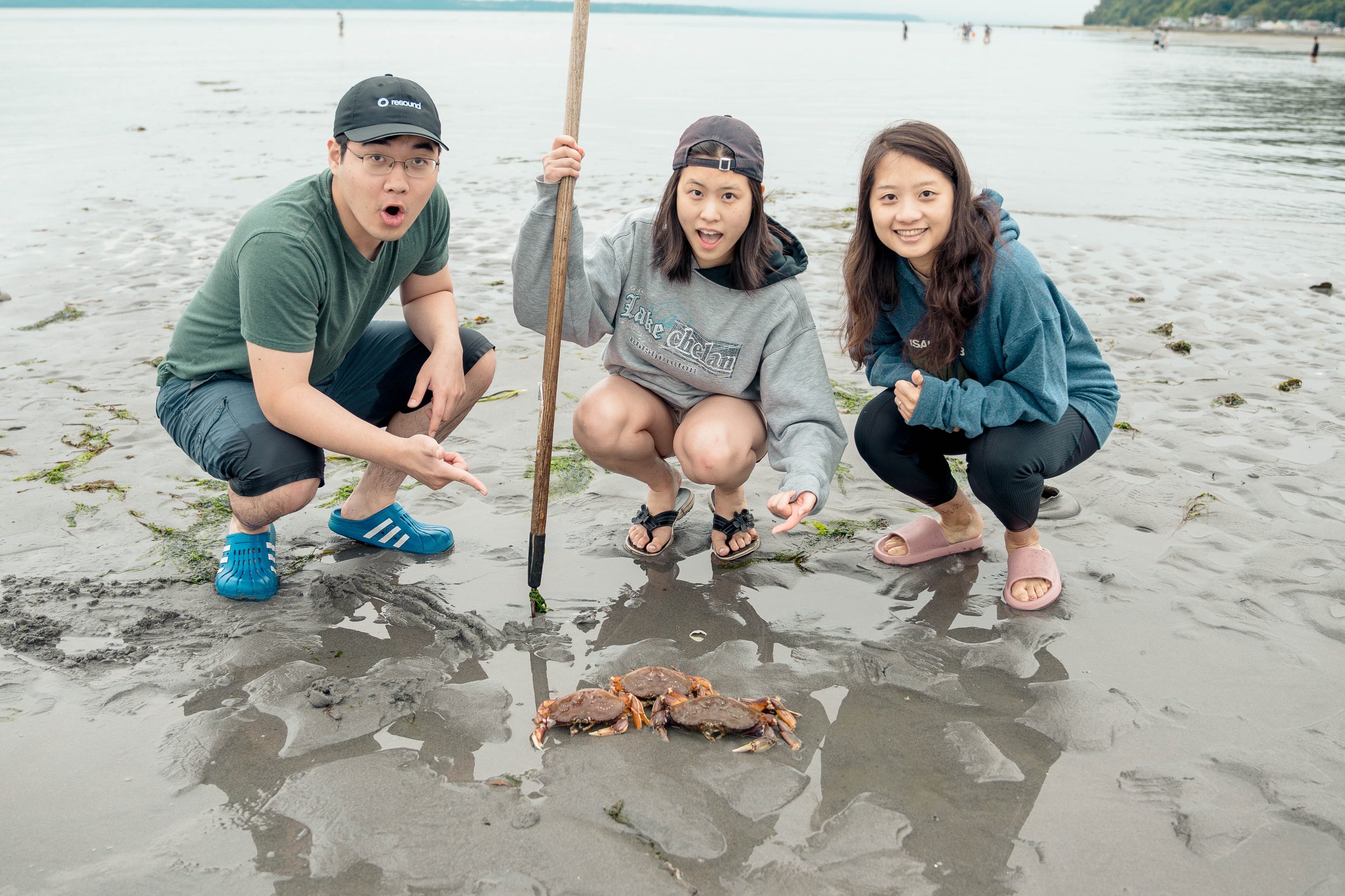 Three people squatting on a sandy beach near a small pile of crabs, holding a crab trap pole, with the ocean background and other beachgoers in the distance.