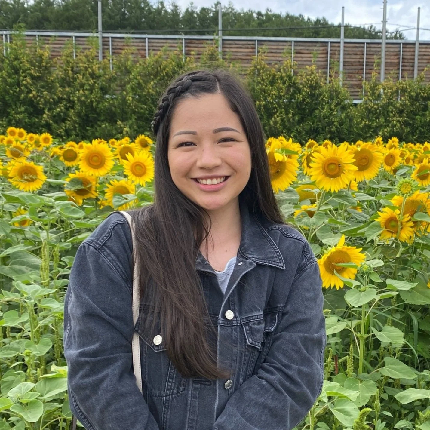 A young woman with long dark hair, wearing a black denim jacket, stands in a field of blooming sunflowers, smiling at the camera.