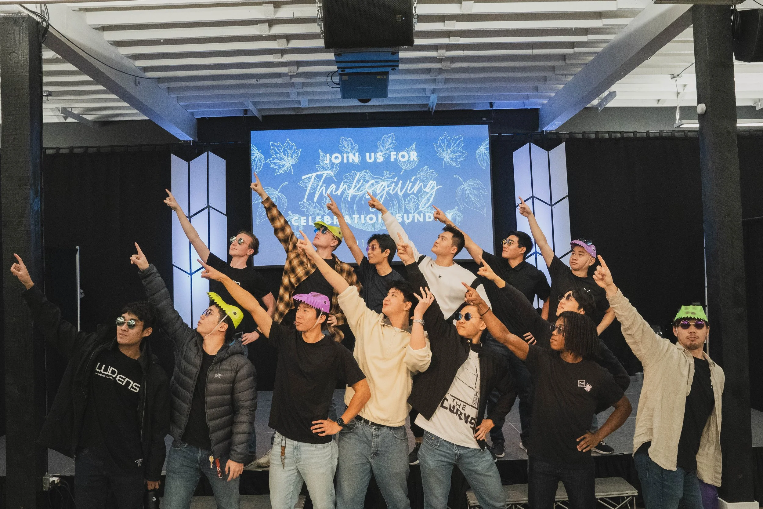 Group of people on stage at a Thanksgiving celebration, posing with arms extended and pointing upwards, some wearing sunglasses and colorful hats. A screen behind them displays the message 'Join us for Thanksgiving Celebration.'