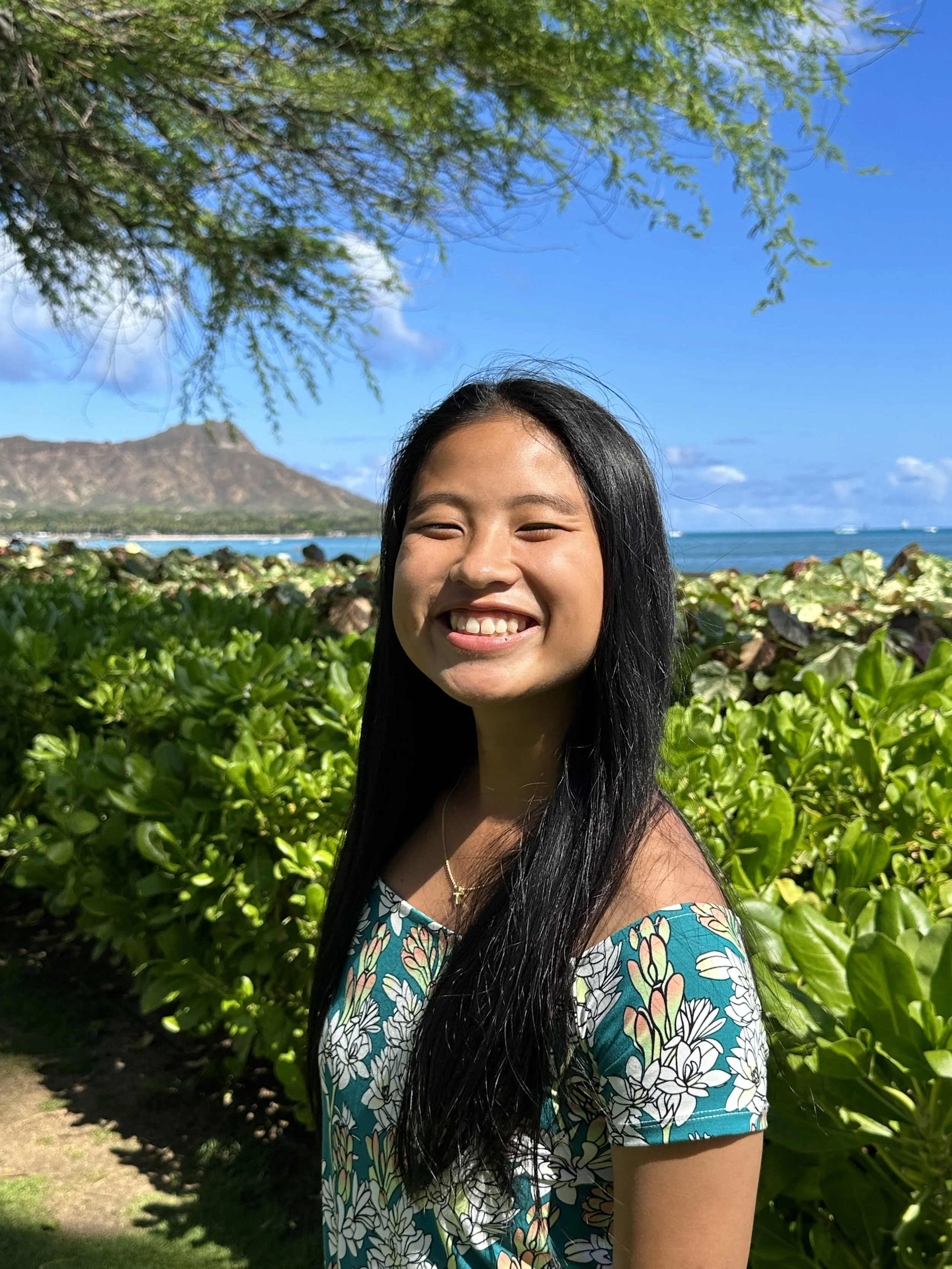 A young woman with long black hair, smiling in a floral dress, standing outdoors near green bushes with a mountain, ocean, and blue sky in the background.