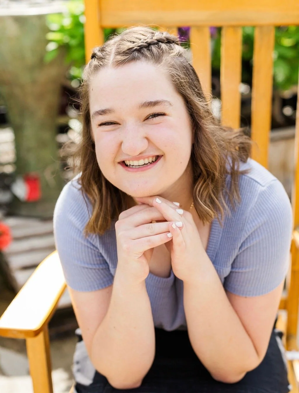 A smiling young woman with light brown hair in a braid, sitting outdoors on a wooden chair, with green foliage in the background.