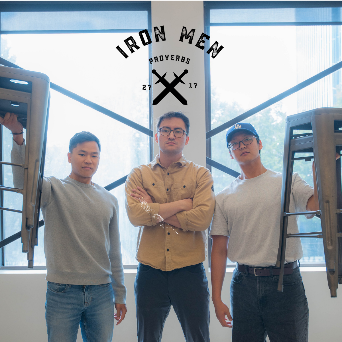 Three men standing in front of large windows with sunlight. The man in the middle has arms crossed, wearing glasses. The man on the left is wearing a gray sweatshirt, and the man on the right is wearing a gray T-shirt, a blue cap, and glasses. Two chairs are held up on each side of the group. The logo 'Iron Men Proverbs 27:17' is displayed above them.